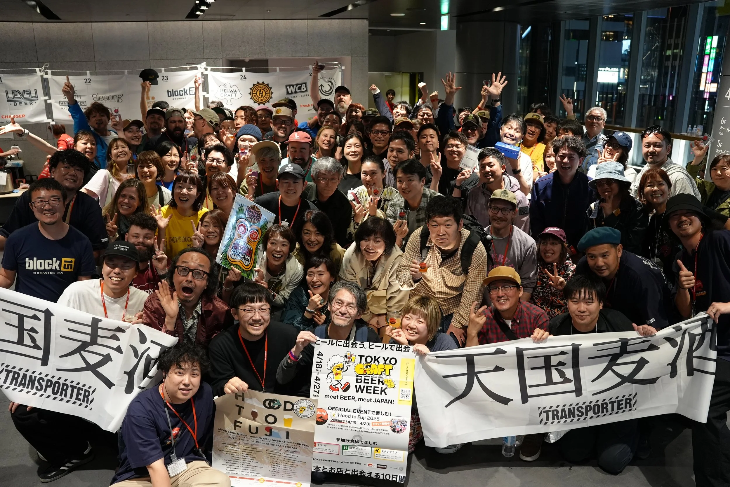 A large group of people gathered indoors for a photo at Tokyo Craft Beer Week, holding banners and signs, some making peace signs, with bright smiles.