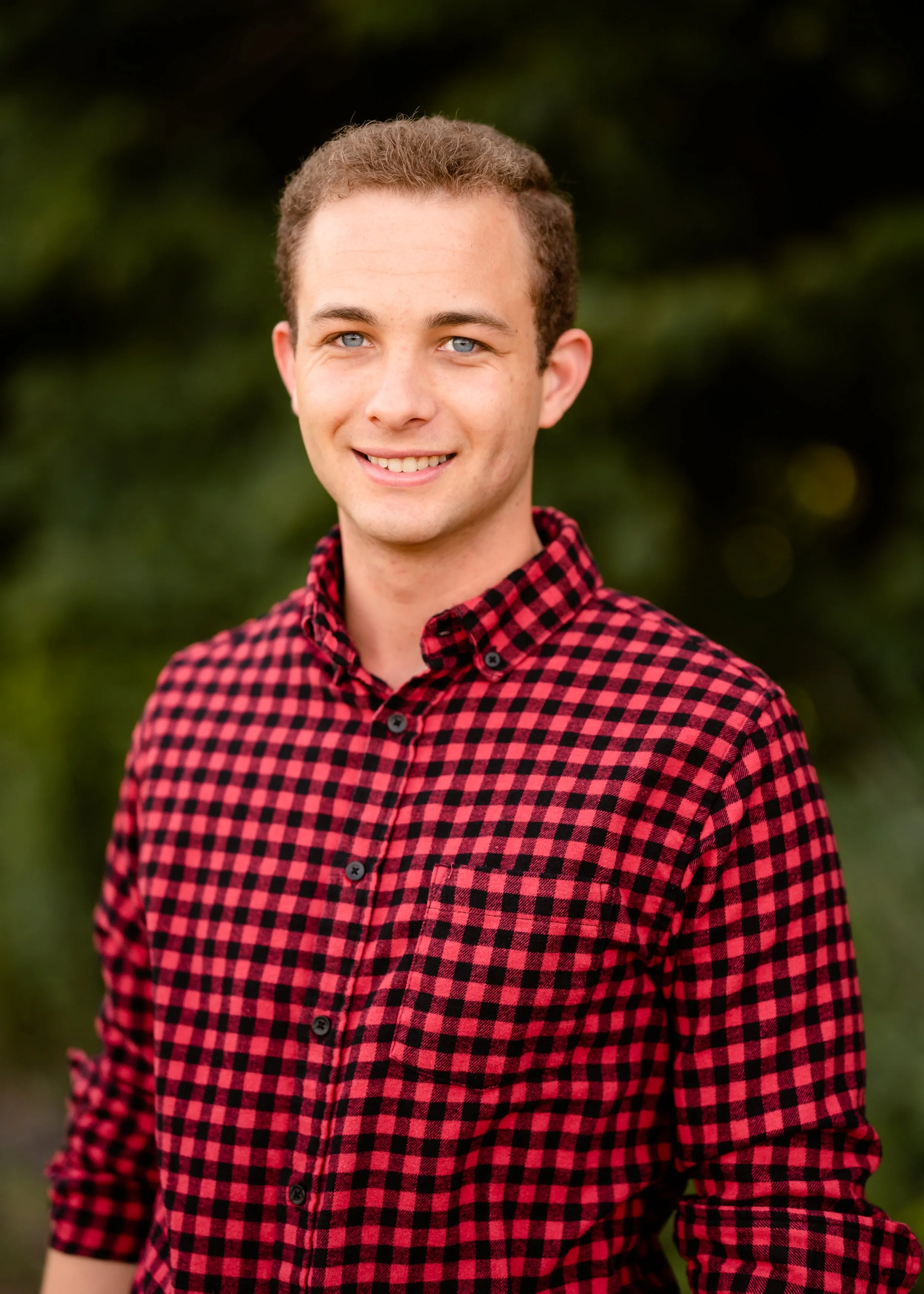 Young man with short curly hair, blue eyes, and a friendly smile, wearing a red and black checkered button-up shirt, standing outdoors with greenery in the background. Isiaah Winchester. Tessitura.