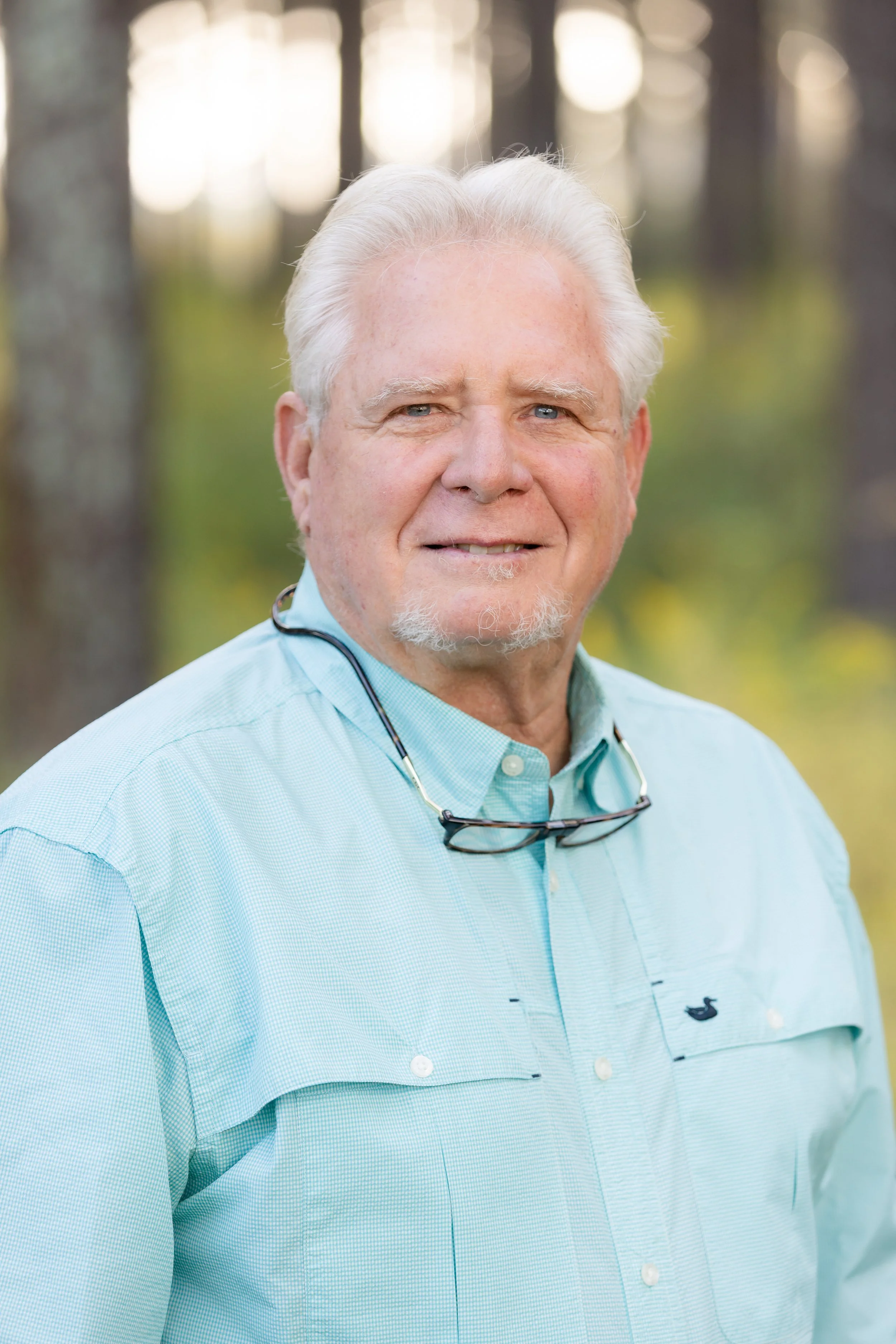 Greene Forest & Farm founder smiles outdoors, wearing a light blue button-up shirt with glasses hanging around his neck, and a forest background.
