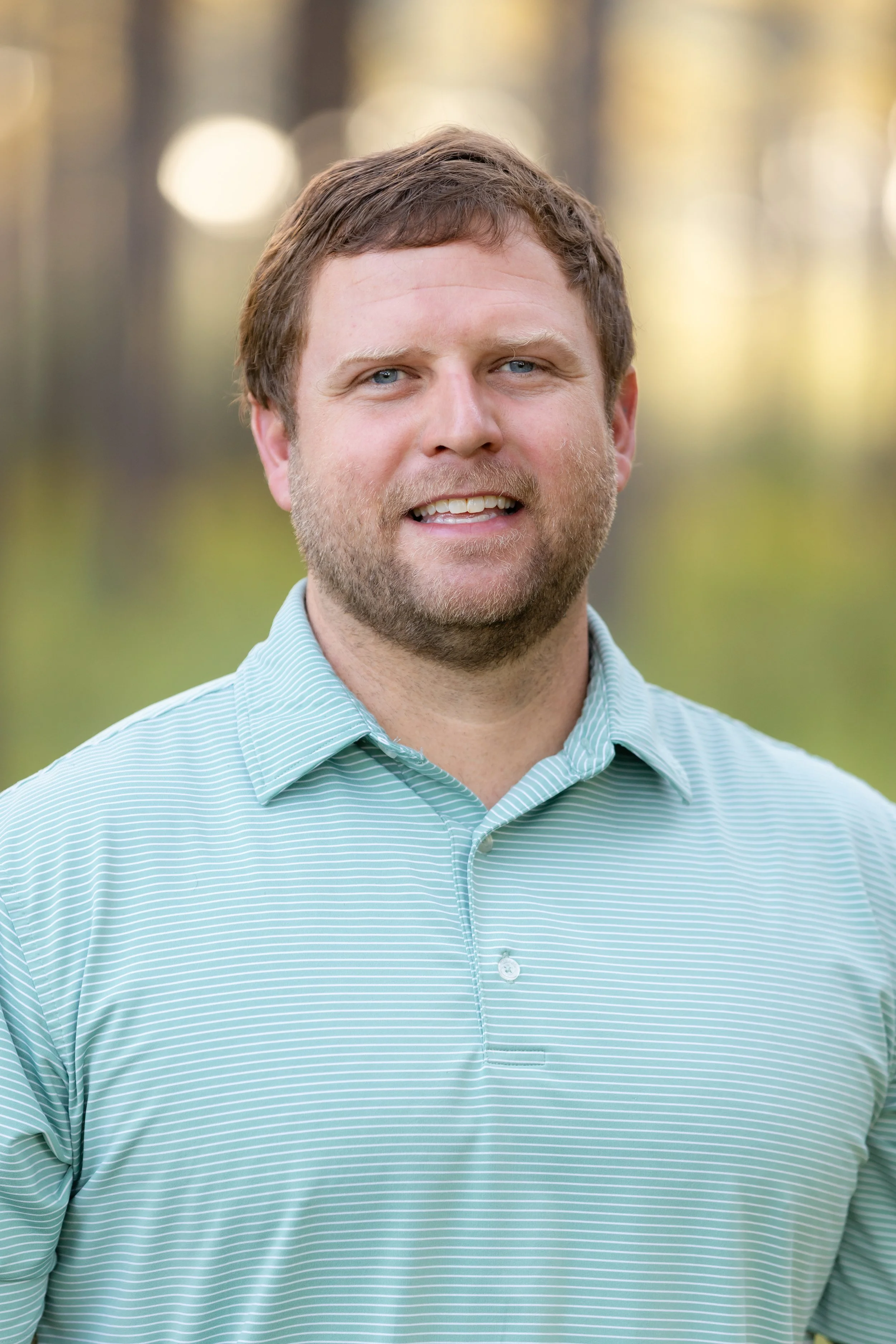 A man with brown hair, blue eyes, and a beard, wearing a light blue and white striped polo shirt, standing outdoors with a blurred background of trees and sunlight.