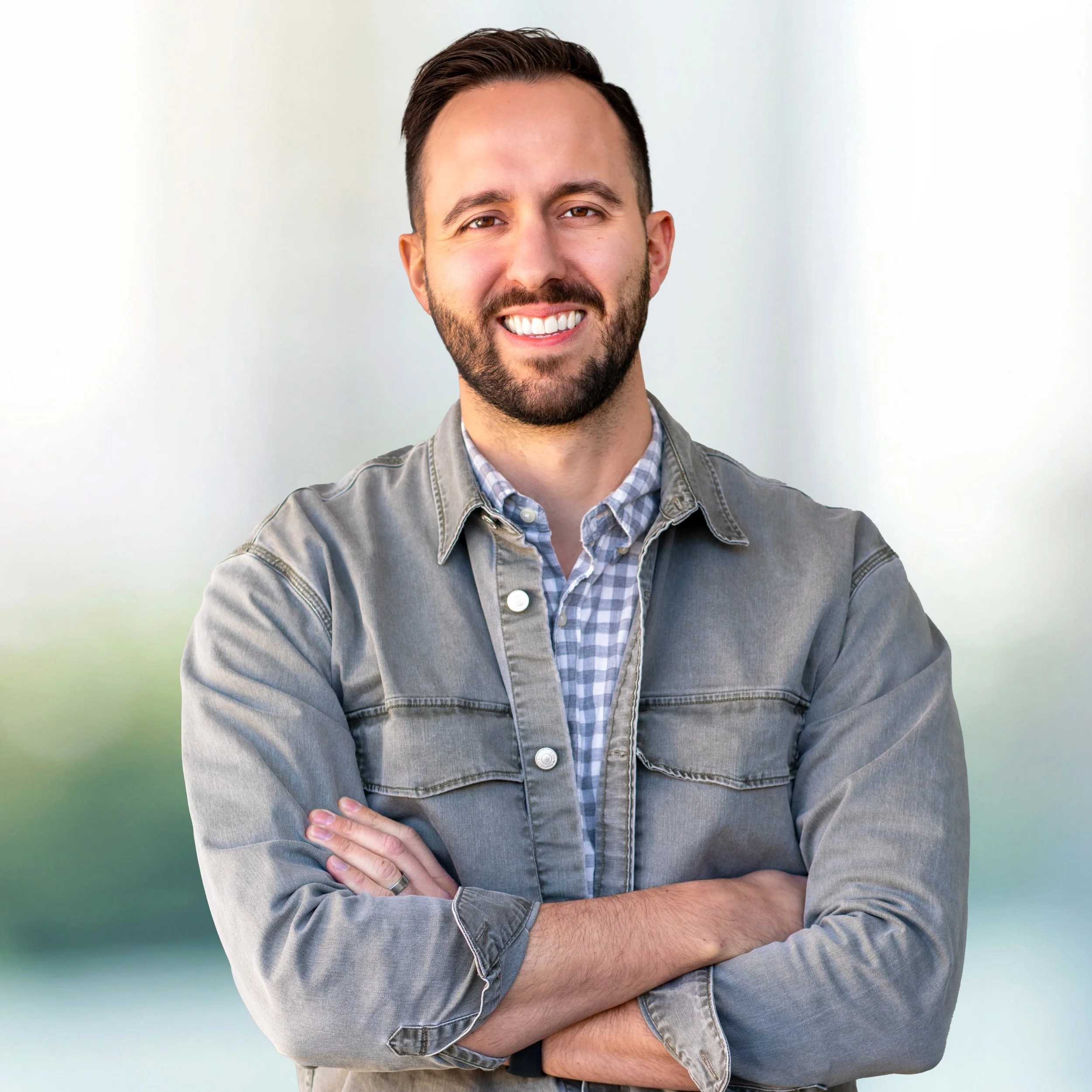 A smiling man with short dark hair and a beard, wearing a gray denim jacket over a checkered shirt, standing against a blurred background with shades of green and white.