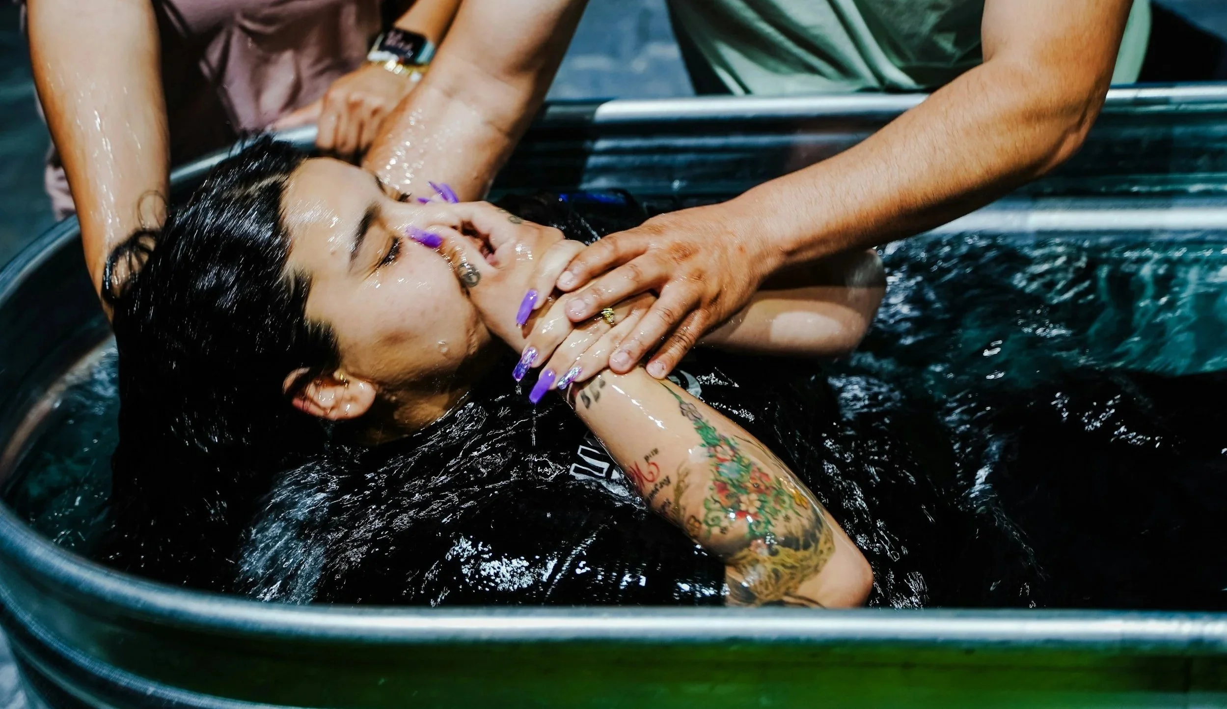 A woman with dark, wet hair receives a baptism in a large metal basin, with water being poured over her head by a person whose hands are on her face and neck during a religious baptism ceremony.