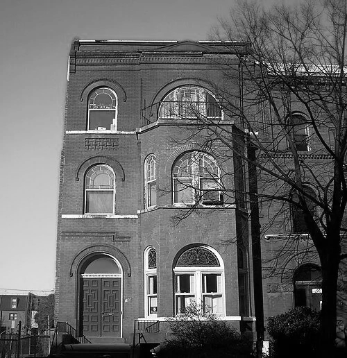 Black and white photo of a multi-story brick building with arched windows and a front door, a leafless tree in front, and a clear sky in the background.
