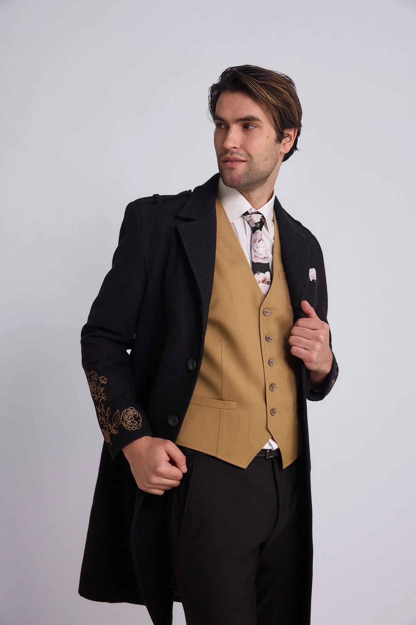 Man wearing a black coat with floral embroidery on the sleeves, a beige waistcoat, a white shirt, a floral tie, and black pants, posing against a plain light gray background.
