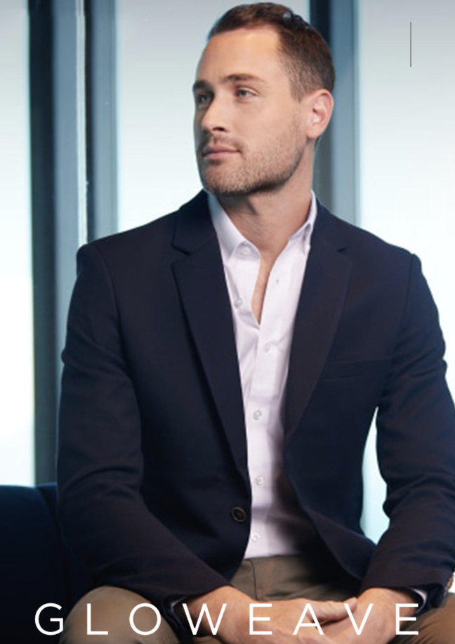 A man in a dark suit and white shirt sitting in a modern office, looking to the side with a thoughtful expression.