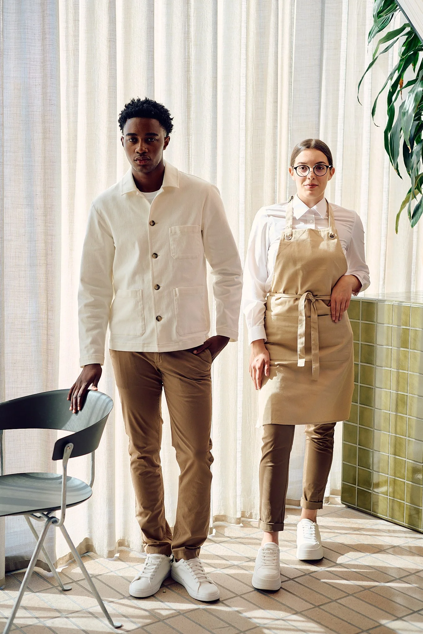 Two individuals, one male and one female, standing indoors near cream-colored curtains. The male wears a white jacket and khaki pants with white sneakers. The female wears a white shirt, beige apron, matching khaki pants, and white sneakers. There is