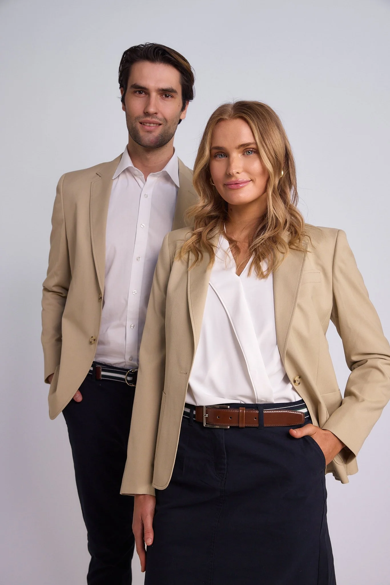 A man and woman dressed in business casual attire standing against a plain gray backdrop. The man is in the background, wearing a beige blazer, white shirt, and dark pants. The woman in front is wearing a beige blazer, white blouse, and a dark skirt,