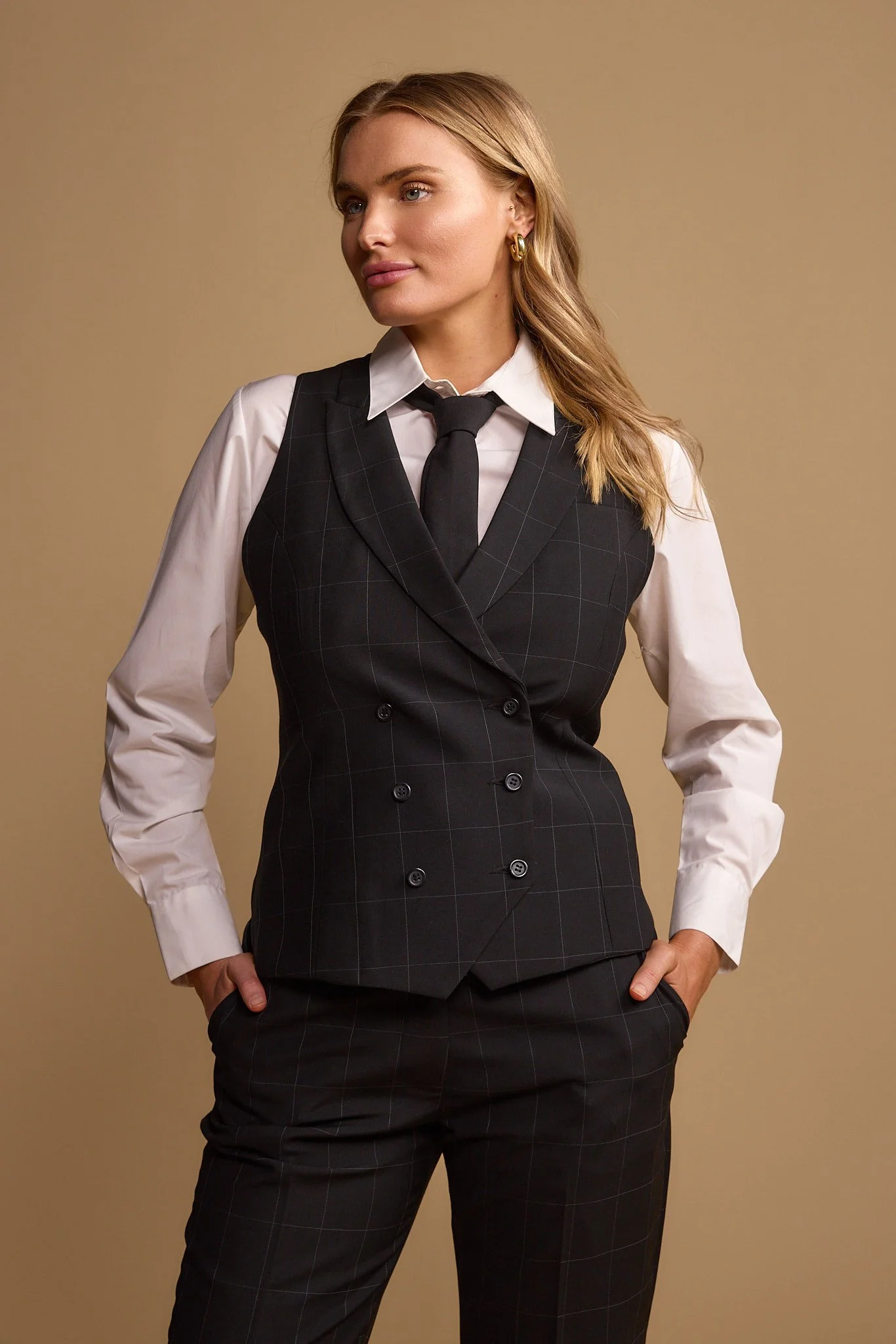 A woman wearing a black plaid double-breasted vest over a white dress shirt paired with matching plaid pants and a black tie, posing with her left hand in her pocket and her right hand at her side against a plain beige background.