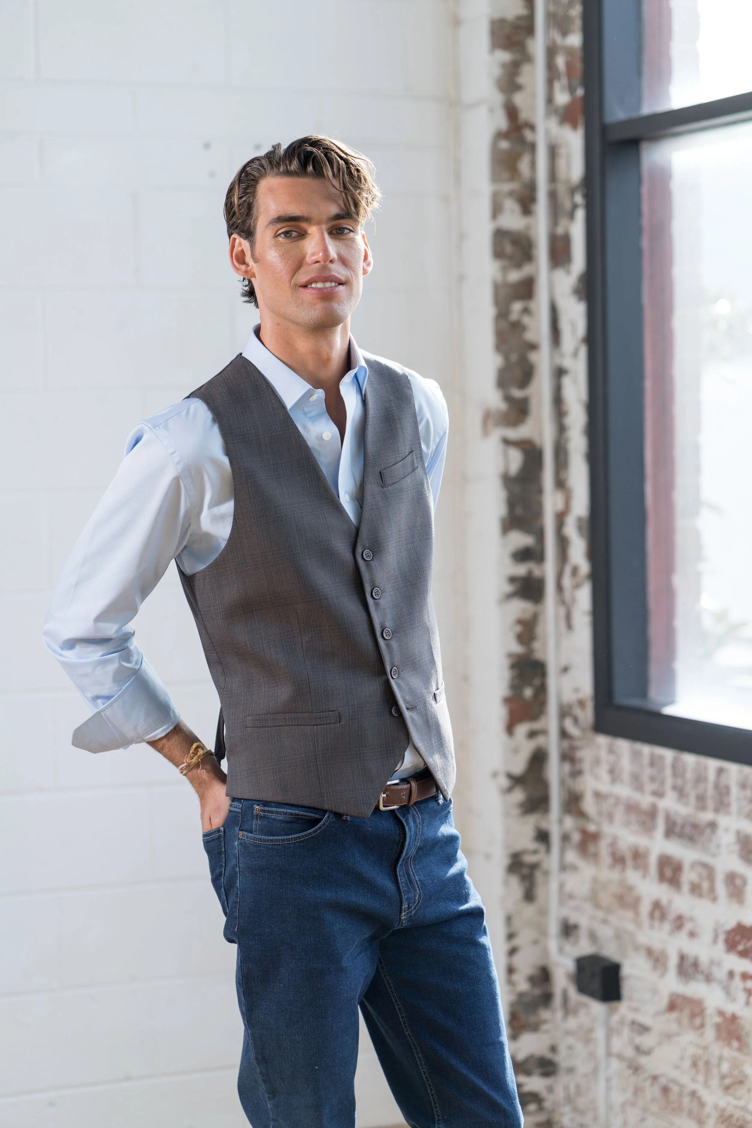 Young man with stylish hair, dressed in a light blue shirt and gray vest, standing inside a modern industrial-style space with exposed brick wall and large window.