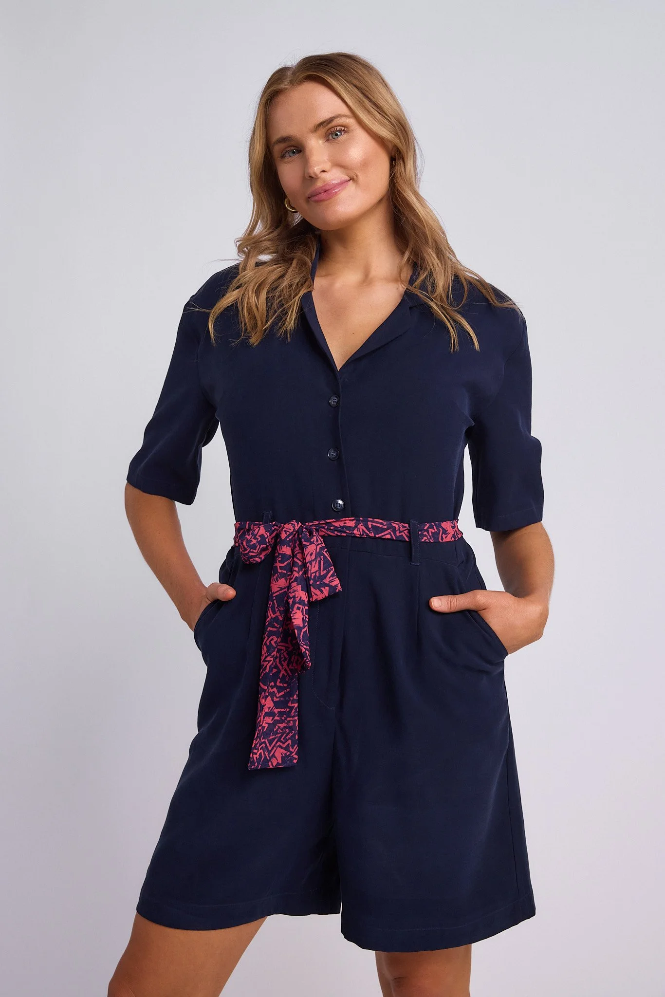 A woman with wavy, shoulder-length blonde hair wearing a navy blue jumpsuit with rolled-up sleeves, a pink patterned belt, and her hands in her pockets, standing against a light gray background.
