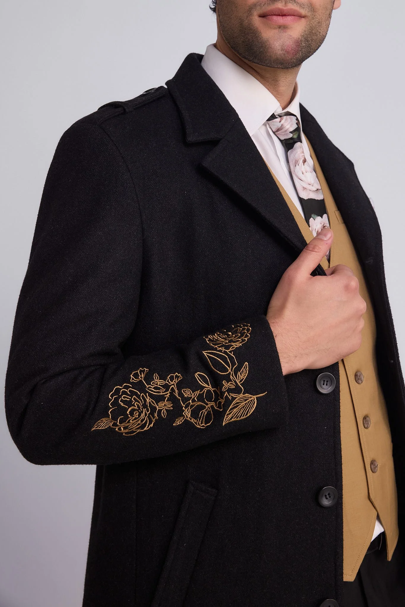 Close-up of a man wearing a black coat with floral embroidery on the sleeve, a floral tie, and a tan vest, pointing to his tie.