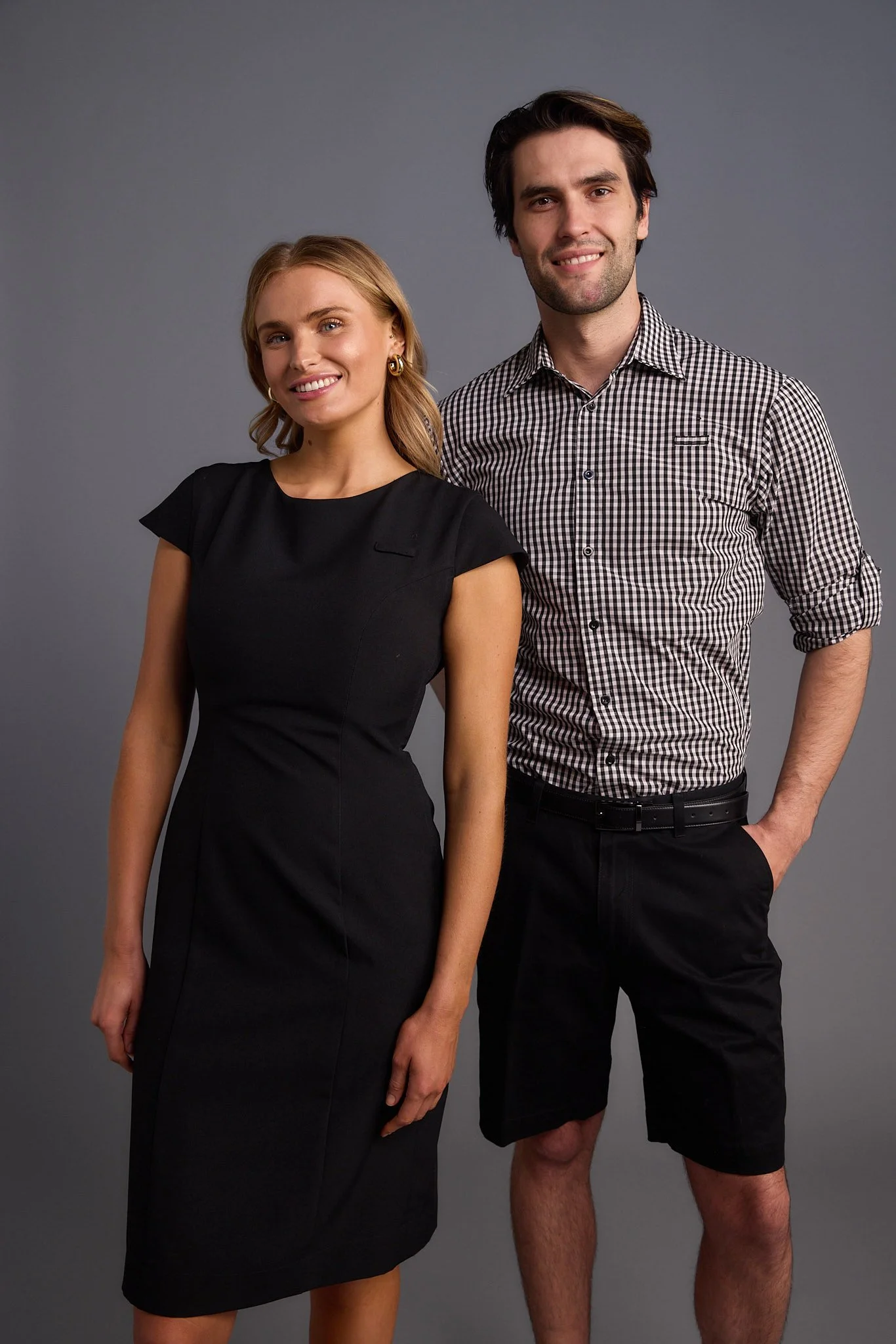 A man and woman standing side by side against a gray background. The woman is wearing a black dress with short sleeves and has blonde hair. The man is wearing a black and white checkered shirt with rolled-up sleeves and black shorts.