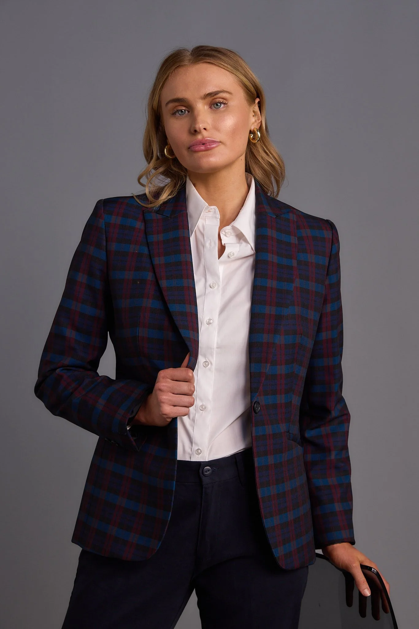 A woman with blonde hair, wearing a checkered blazer over a white shirt, holding a mobile phone, standing against a plain gray background.