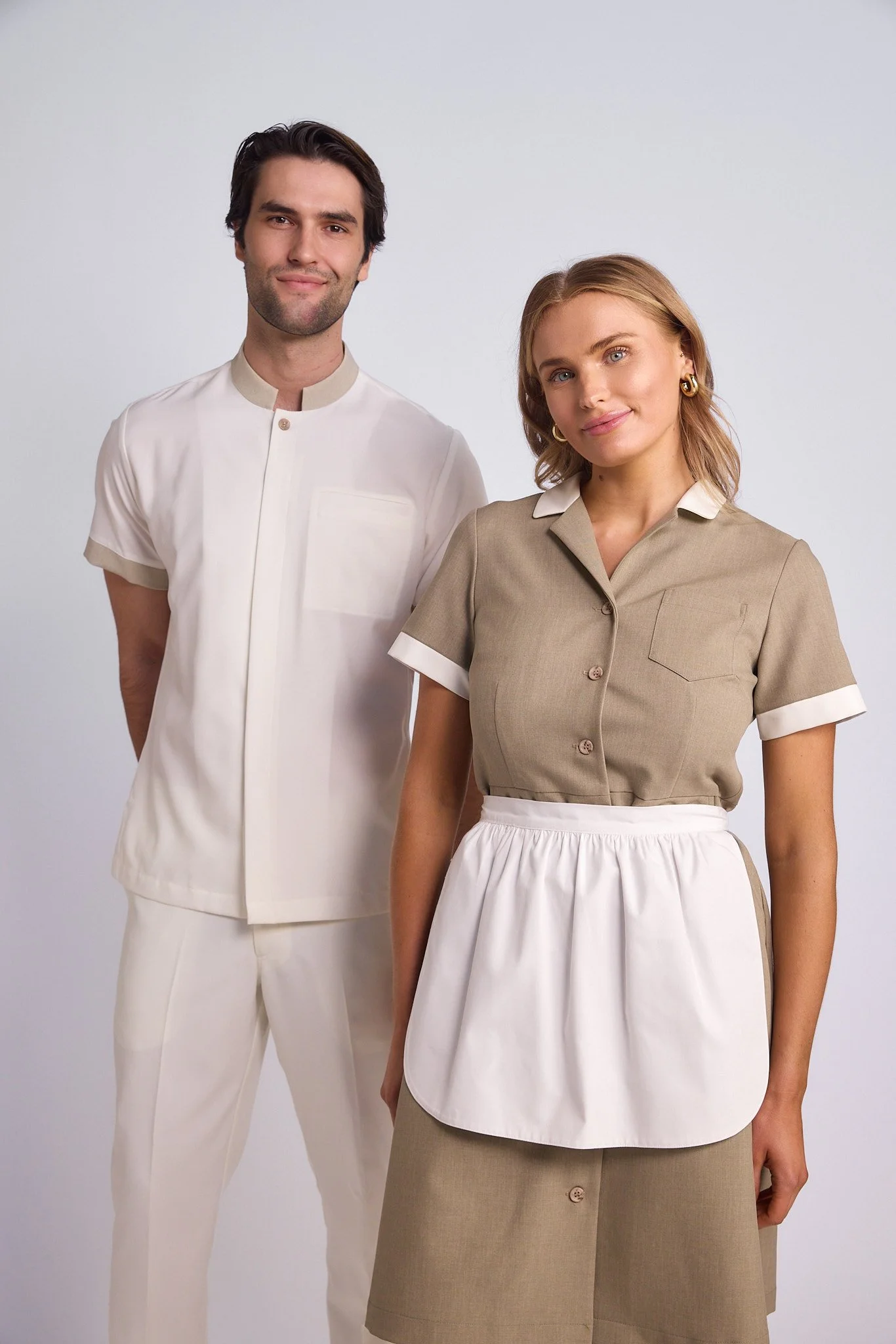A man and a woman standing side by side, both dressed in neutral-colored work uniforms against a plain white background.