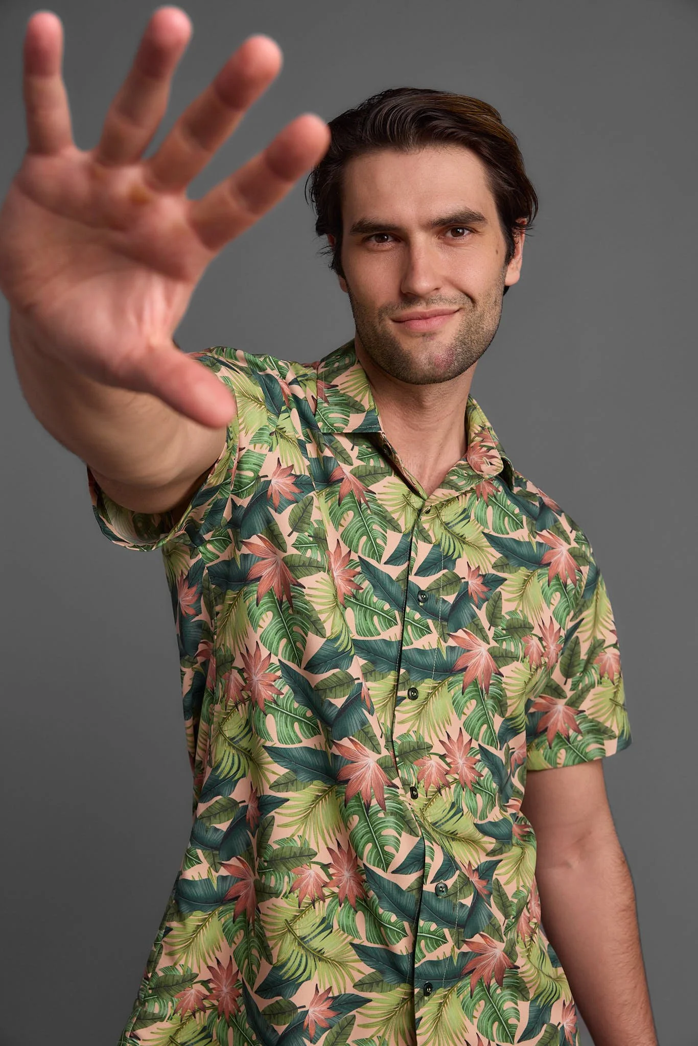 A man with dark hair wearing a colorful tropical print shirt, extending his hand toward the camera with a neutral expression and slight smile, standing against a plain gray background.