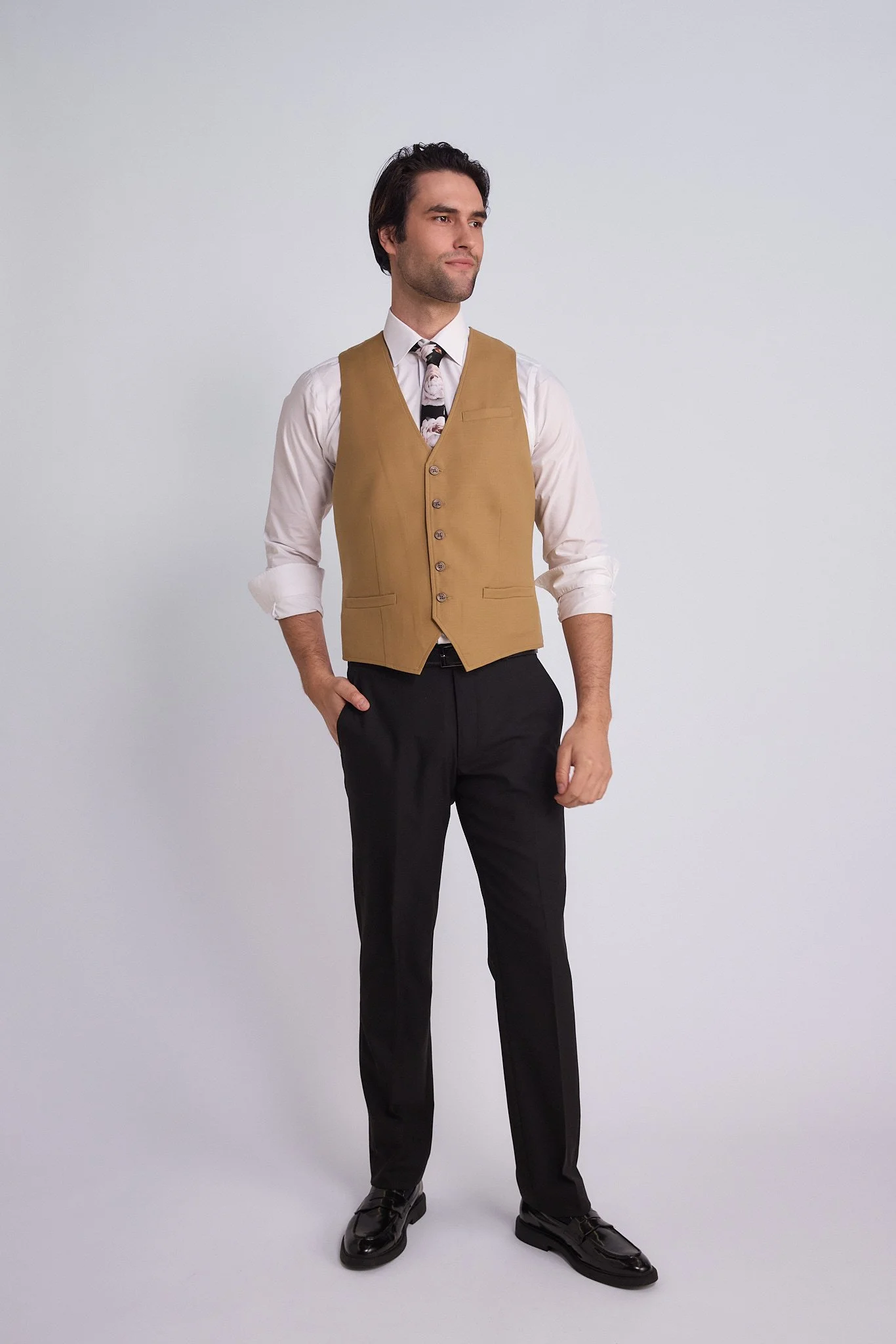A man wearing a white shirt, tan vest, black pants, and black shoes standing against a plain white background.