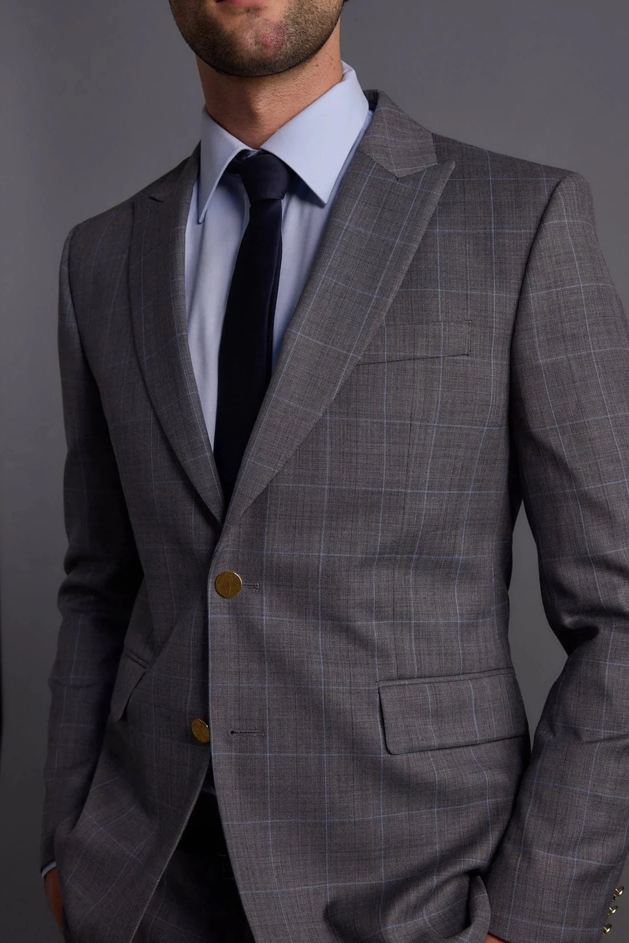 Close-up of a man wearing a fitted gray plaid suit, white dress shirt, and navy tie, against a plain gray background.