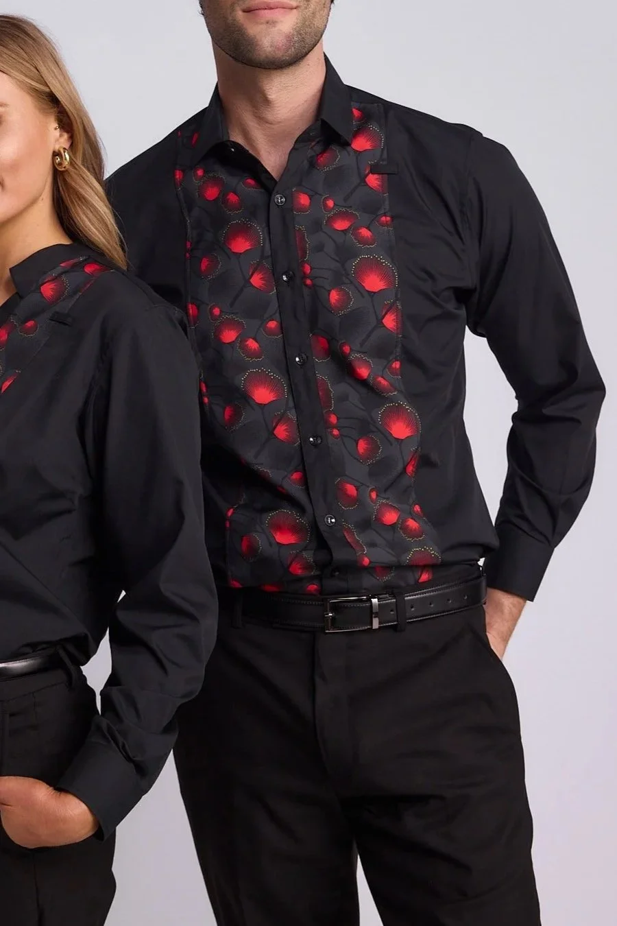 Close-up of a man in a black dress shirt with a red and black patterned front panel, standing with one hand in his pocket, against a plain gray background.