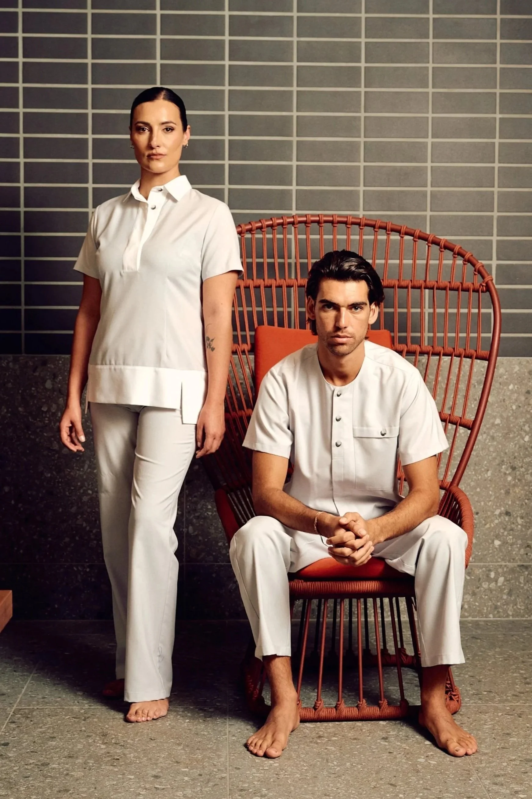 A woman and a man dressed in white, with the woman standing and the man sitting in an orange wicker chair, in front of a gray tiled wall.
