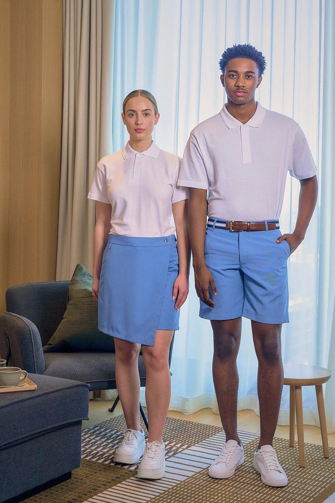 A young man and woman standing in a living room, both wearing matching white collared shirts and light blue bottoms, with the woman in a skirt and the man in shorts.