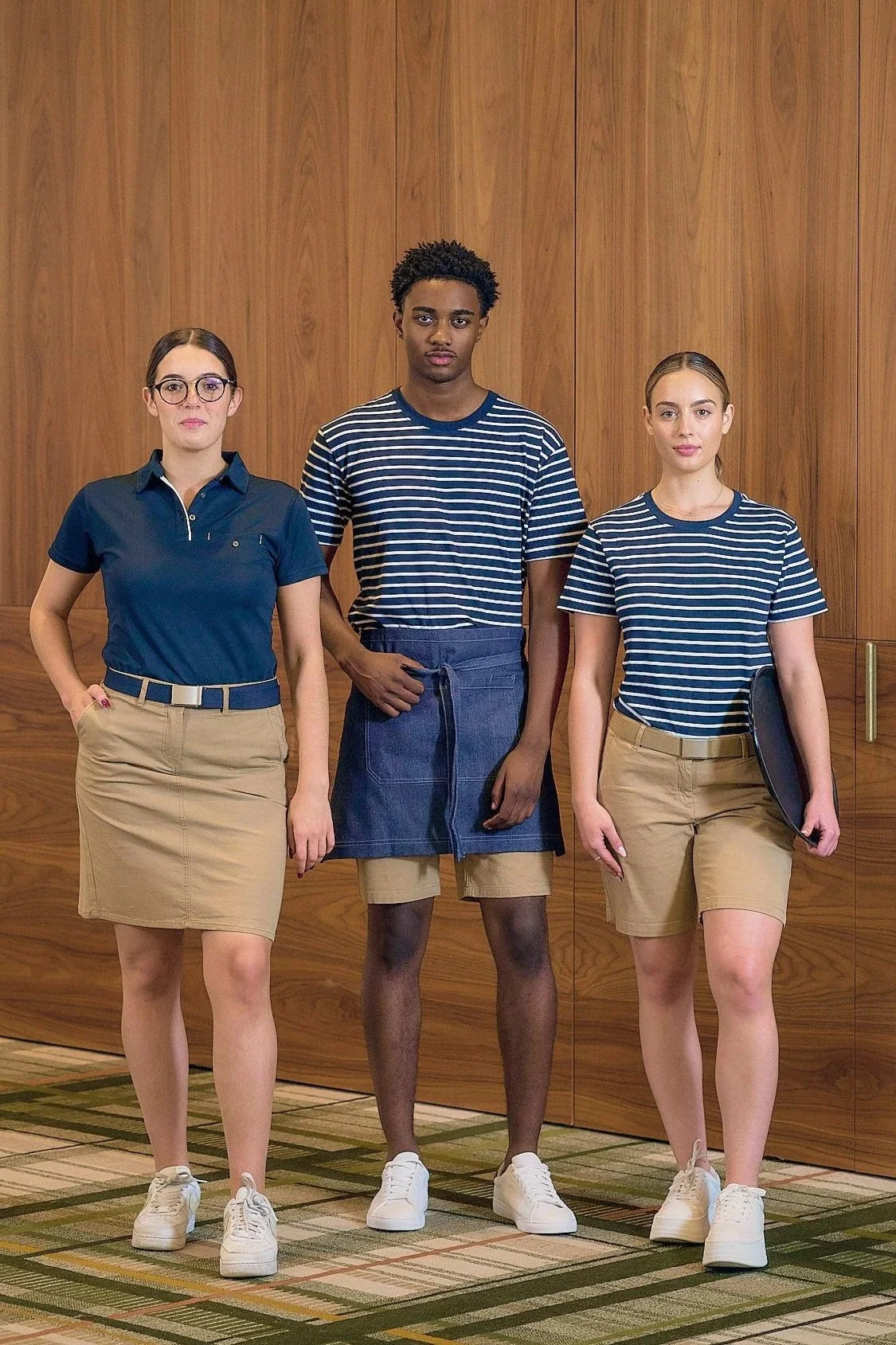 Three young adults stand side by side, posing for a photo indoors against a wooden-paneled wall. The person on the left is a woman with glasses wearing a navy blue polo shirt and beige skirt, the person in the middle is a man wearing a striped t-shir