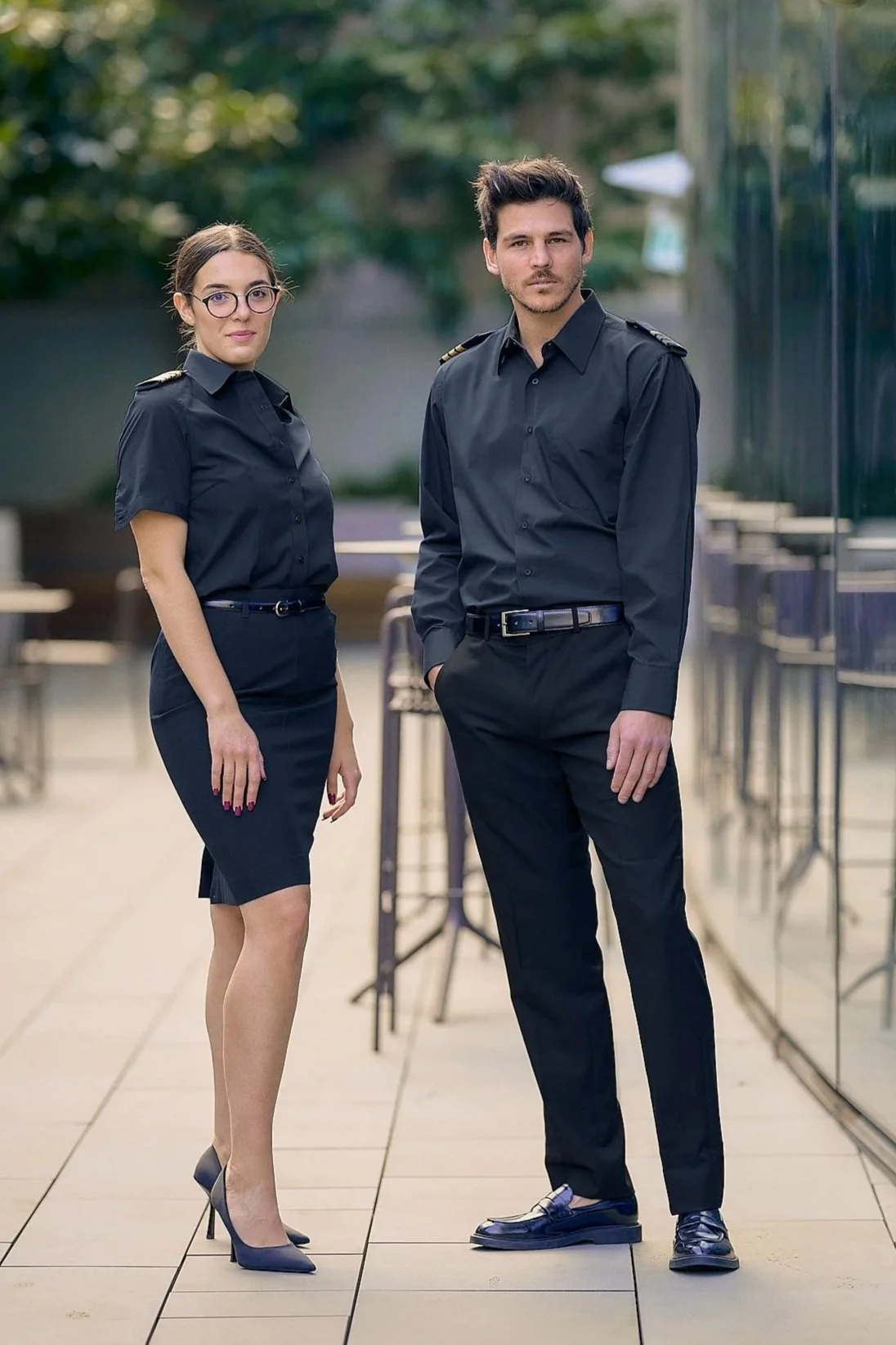 Two individuals, a woman and a man, dressed in black business attire, standing outdoors on a tiled walkway with glass and greenery in the background.