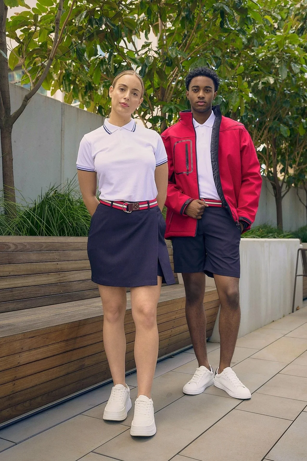 A young woman and young man are standing outdoors in front of a wooden bench and greenery. The woman is wearing a white polo shirt, navy blue skirt, and white sneakers. The man is wearing a red jacket over a white shirt, navy shorts, and white sneake
