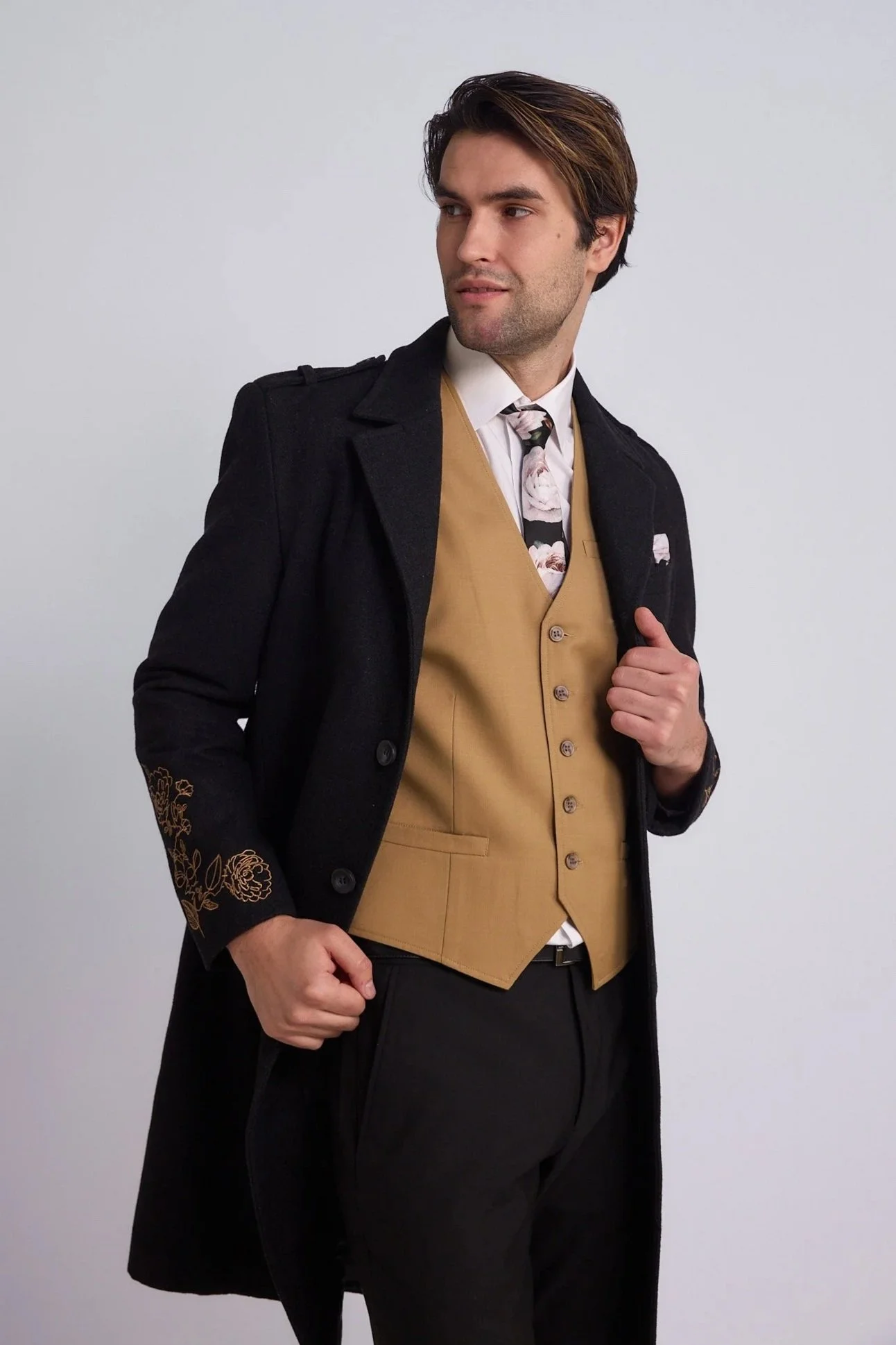 A man wearing a black coat with gold embroidery on the sleeve, a tan vest, a white shirt, and a floral tie poses against a plain white background.
