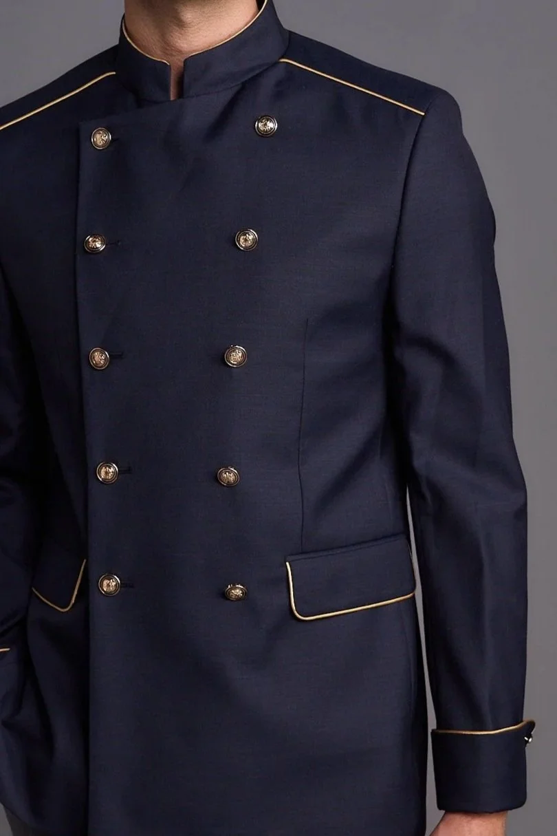 A man wearing a dark navy chef jacket with gold piping and buttons, standing against a plain gray background.
