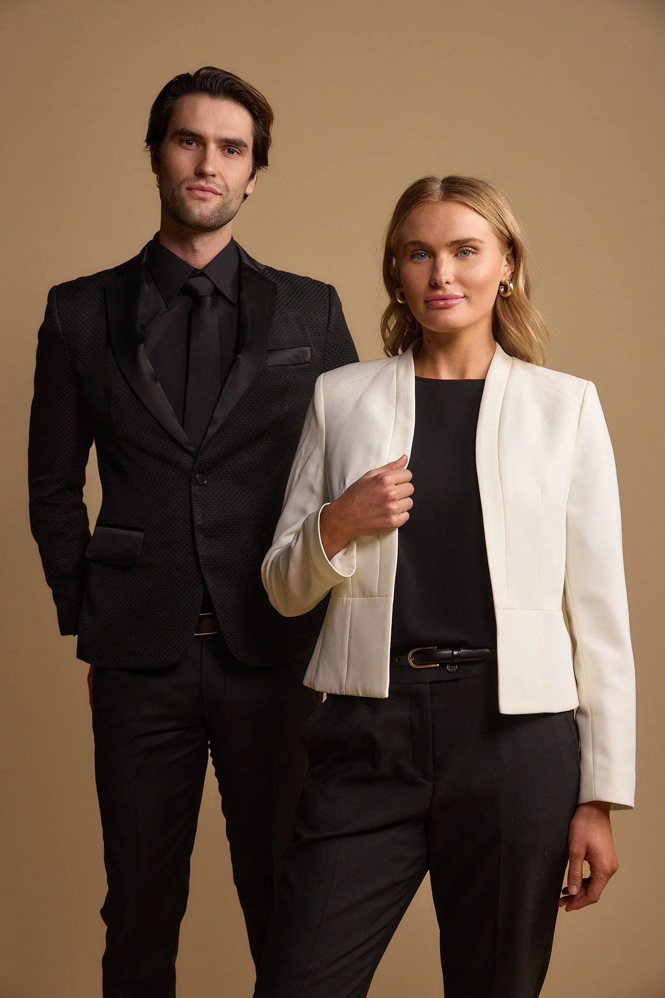 A man and woman standing side by side in formal business attire against a plain beige background.