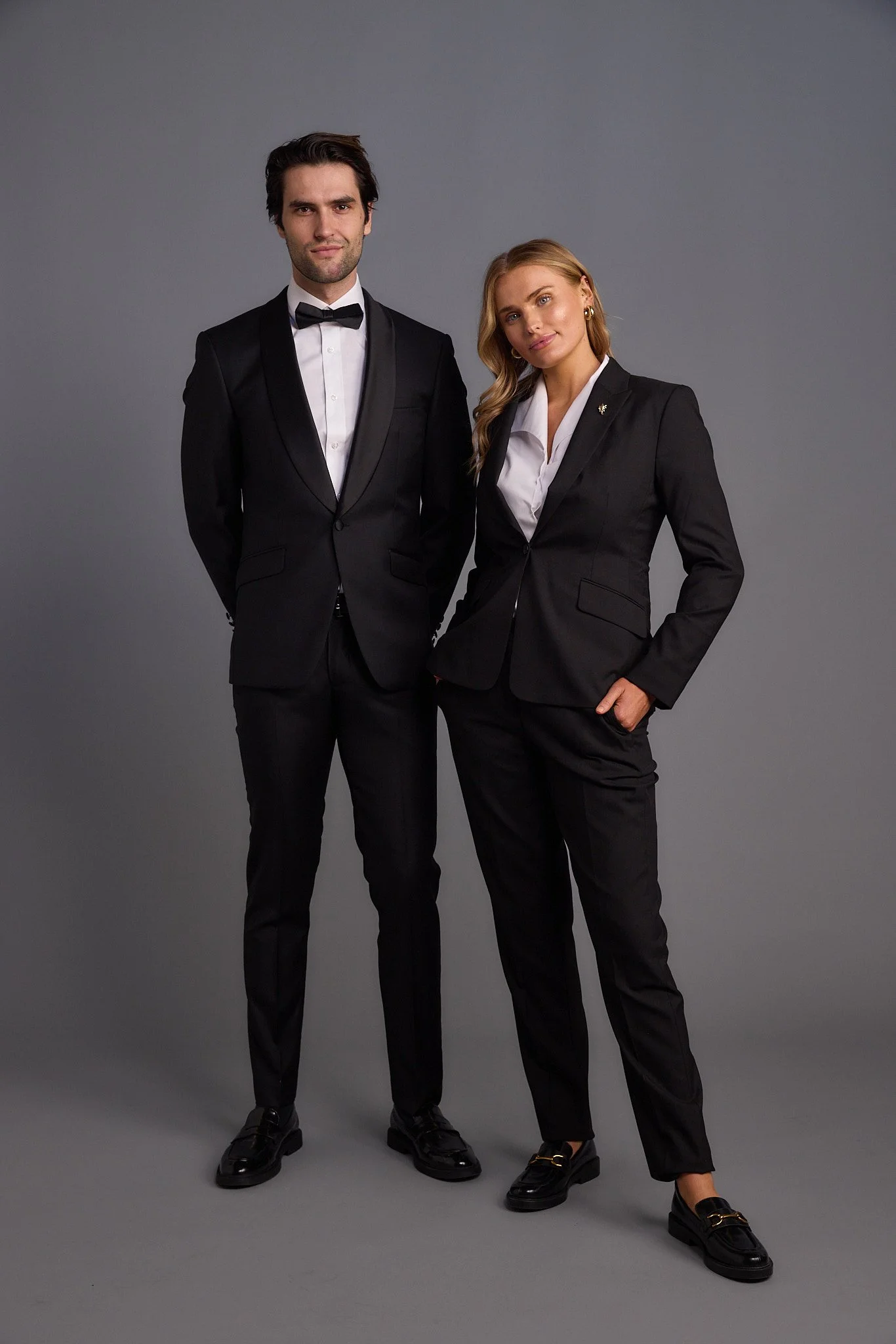 A man and woman dressed in black tuxedos and dress shoes standing against a gray background.