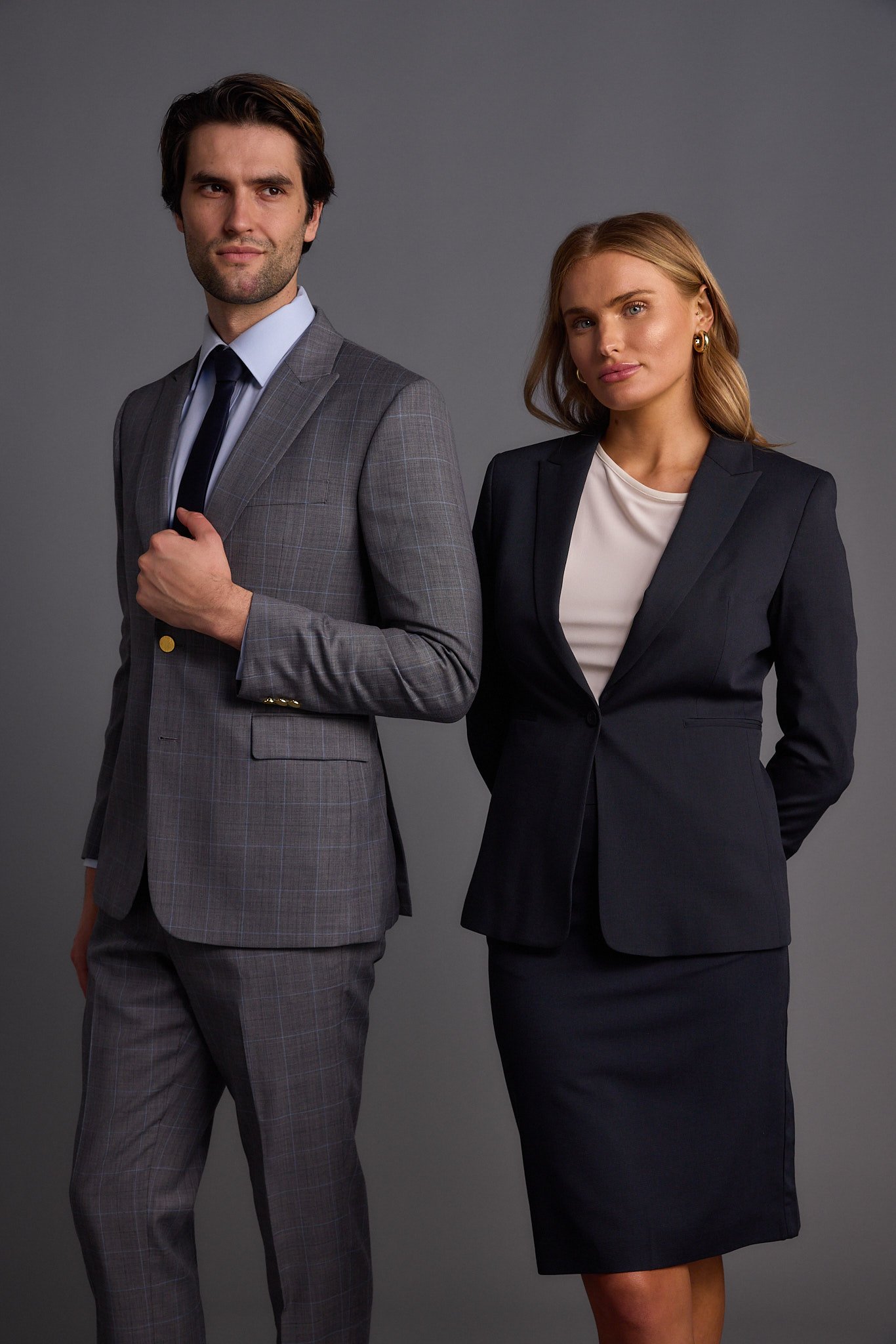 A man and woman dressed in professional business attire standing against a gray background.