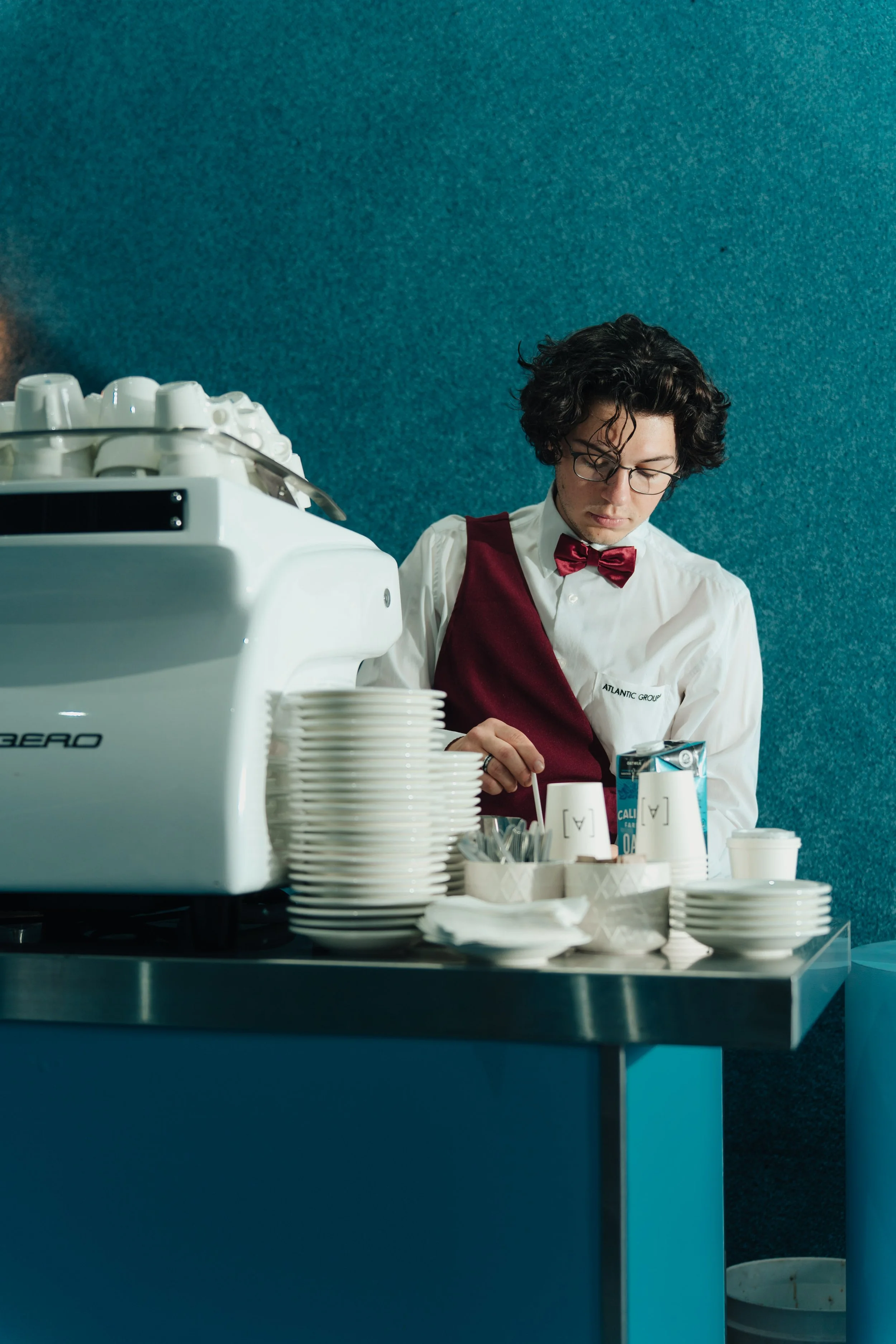 A young man with curly hair and glasses wearing a white shirt, burgundy vest, and bow tie making coffee behind a counter with stacked cups, plates, and a coffee machine.