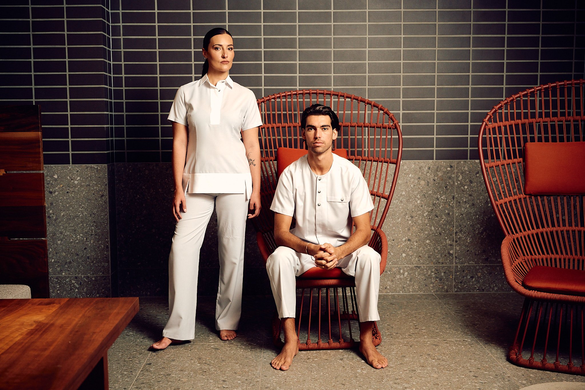 A woman in white medical scrubs standing next to a man seated in a red wicker chair, both in a room with dark gray tiled walls and stone flooring.