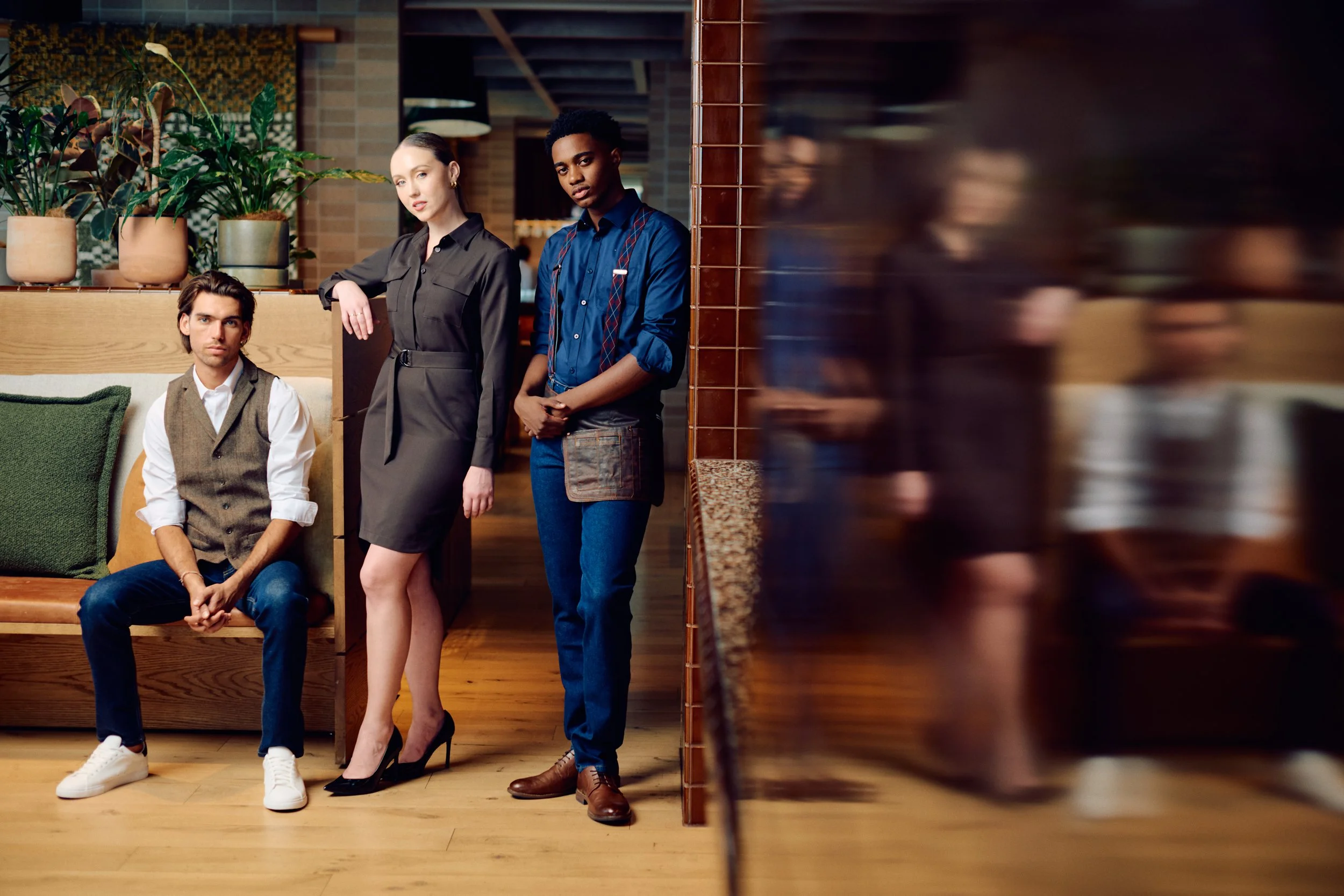 Three young adults in a restaurant, two standing and one sitting, with plants and wooden decor.