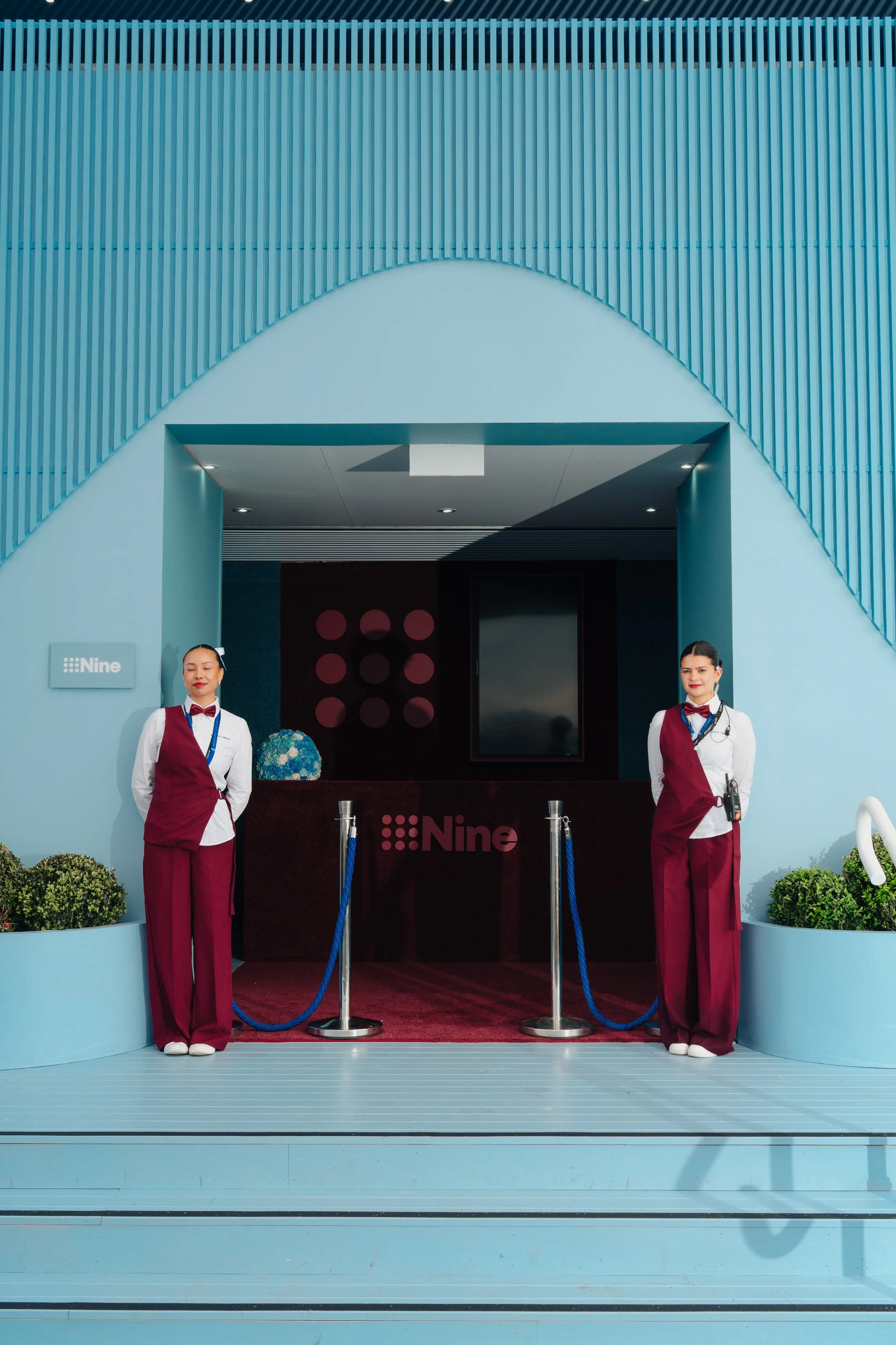 Two women in matching white shirts and maroon vests and pants stand at the entrance of a building with a blue exterior. The building has a sign that says 'Nine' and a red carpet with blue stanchions. The women appear to be staff or greeters.