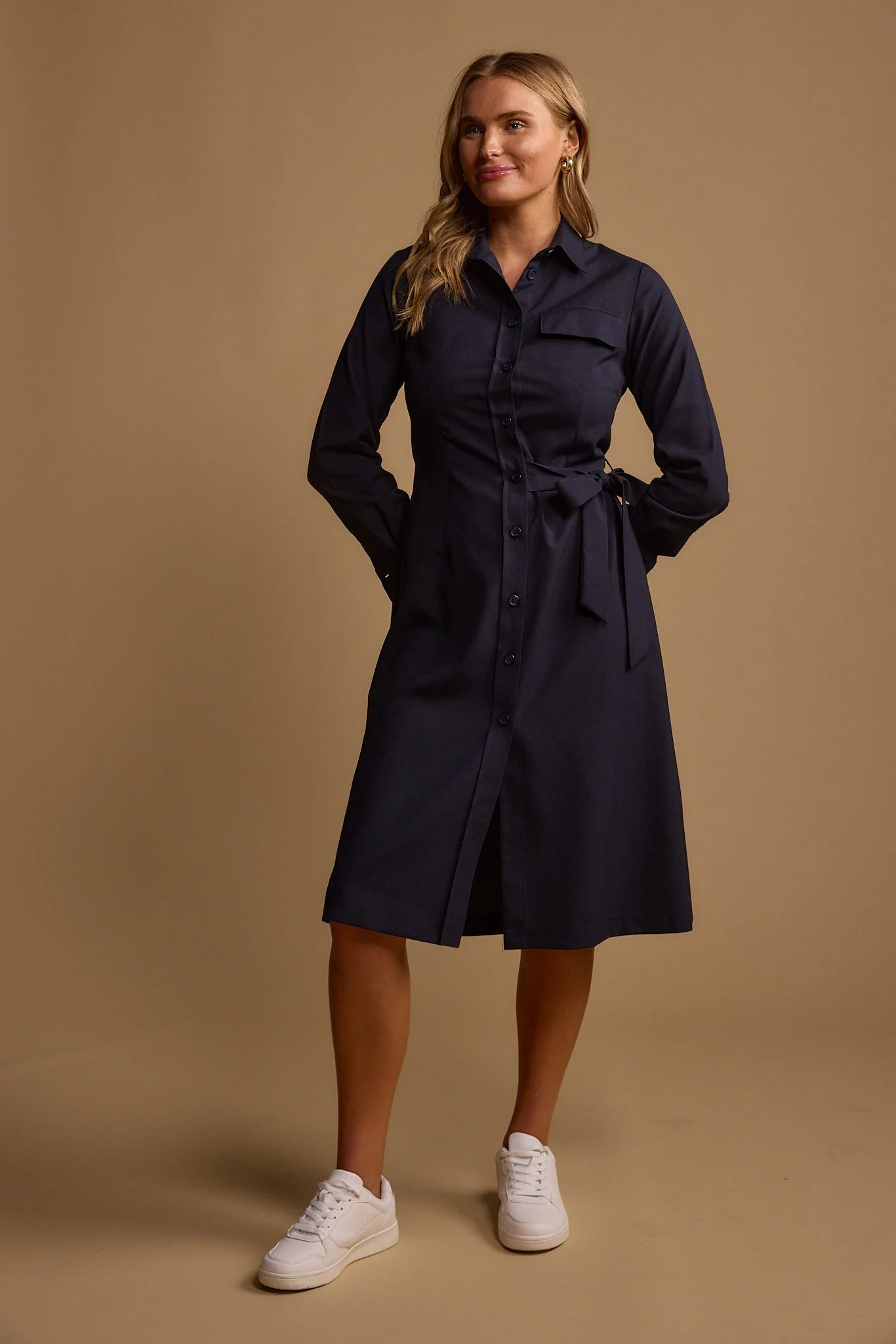 A woman with blonde hair wearing a navy blue shirt dress with a tied waist and white sneakers, standing against a plain beige background.