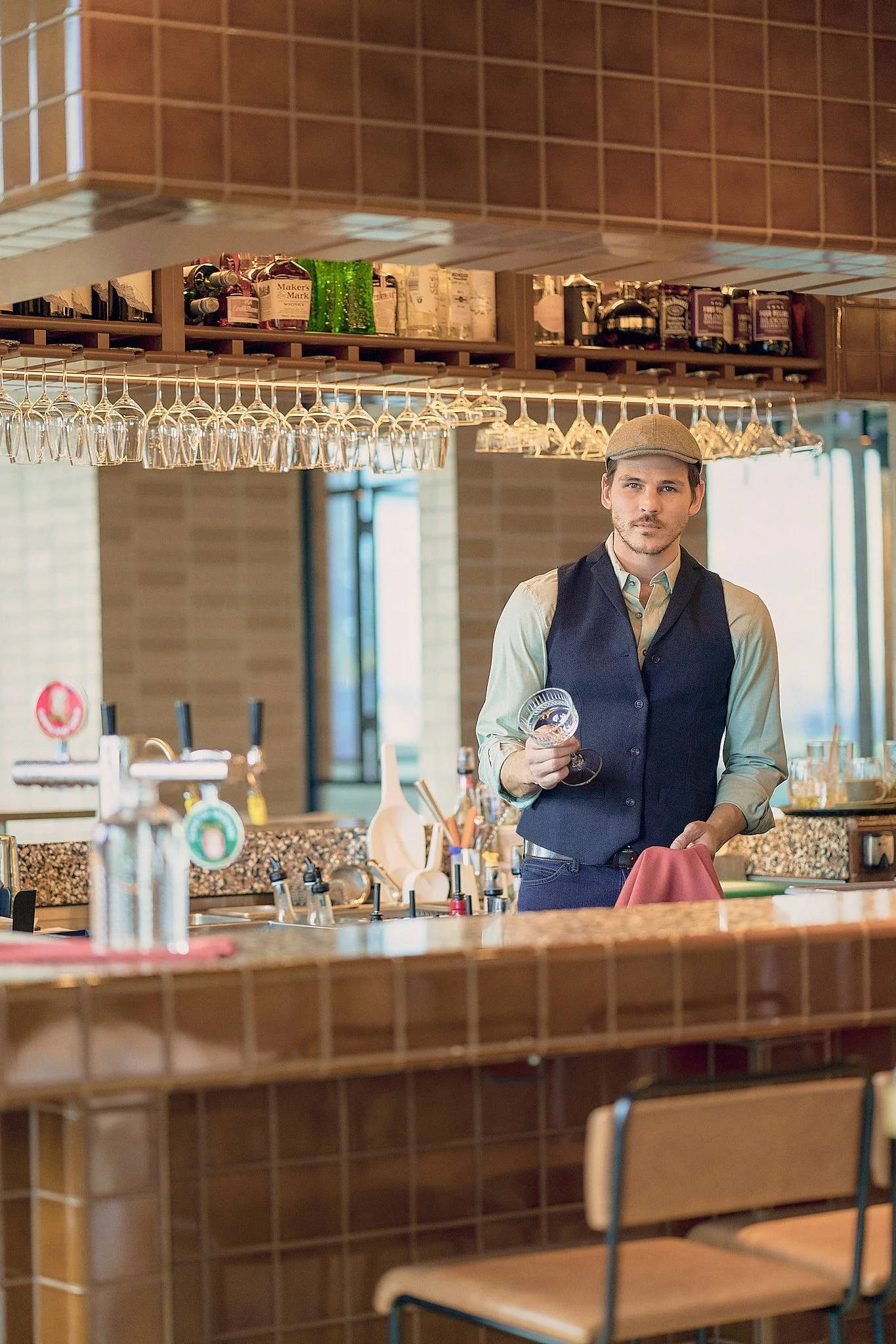 A young male bartender wearing a beige cap, a light-colored shirt, and a dark vest, holding a wine glass and a pink cloth in a bar or restaurant setting.