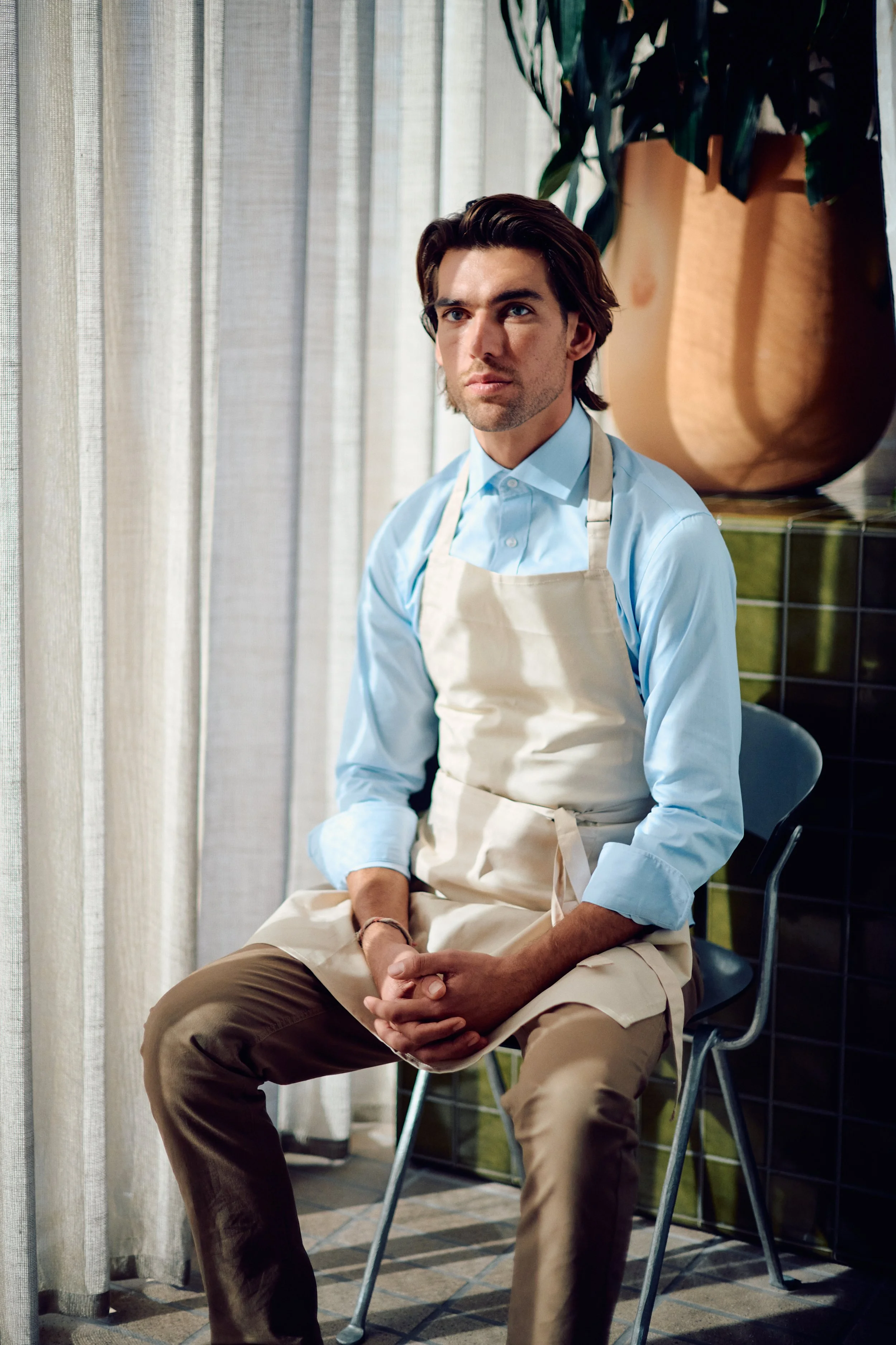 A man with dark hair and blue eyes, wearing a light blue shirt and beige apron, sitting on a chair indoors near a window with beige curtains. Behind him is a large decorative plant in a wooden pot.
