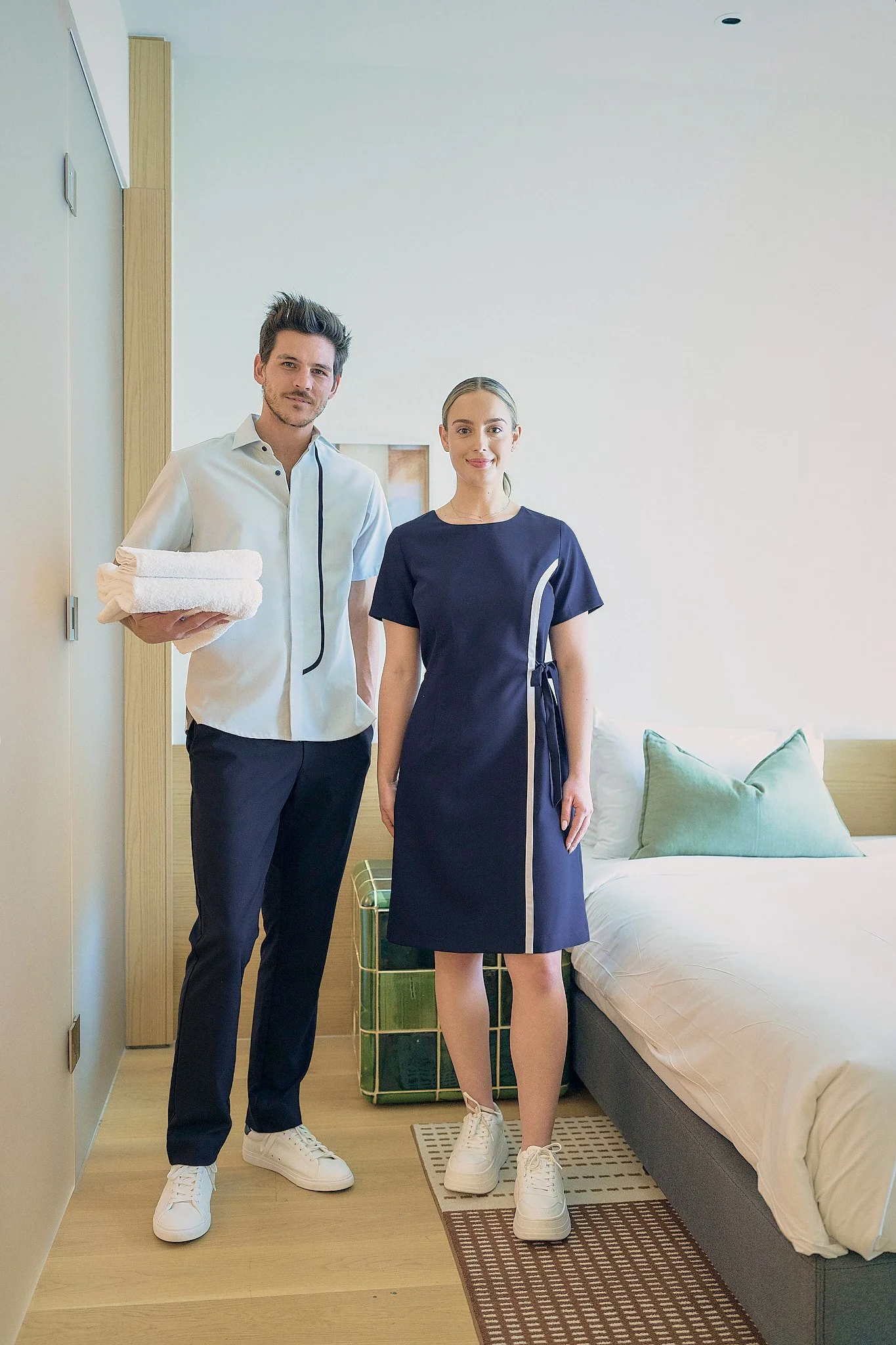A man holding folded towels standing next to a woman in a navy dress inside a modern hotel room.