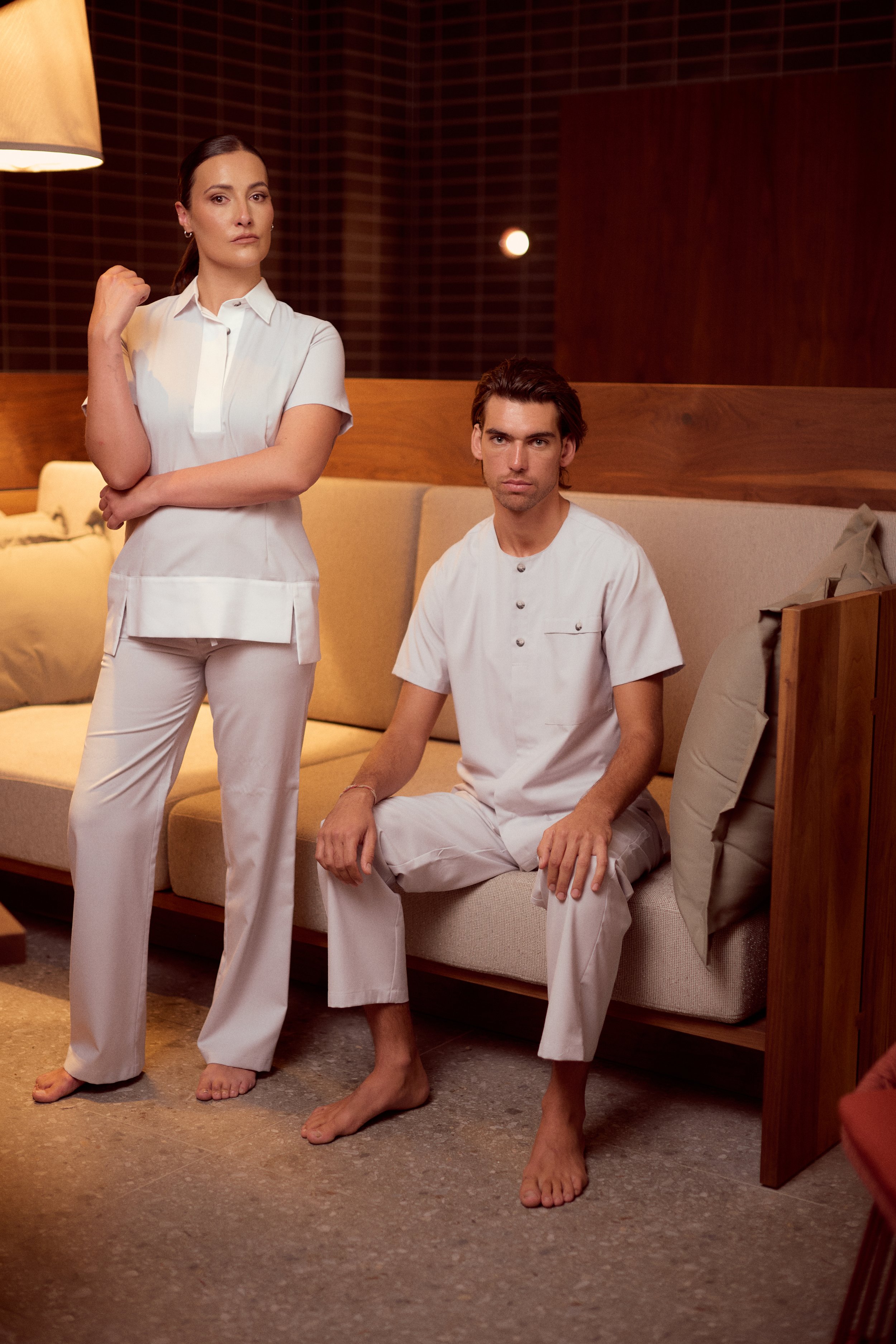 A woman and a man dressed in medical or spa attire sit and stand on a beige sofa in a warmly lit room with wooden walls, both with serious expressions.
