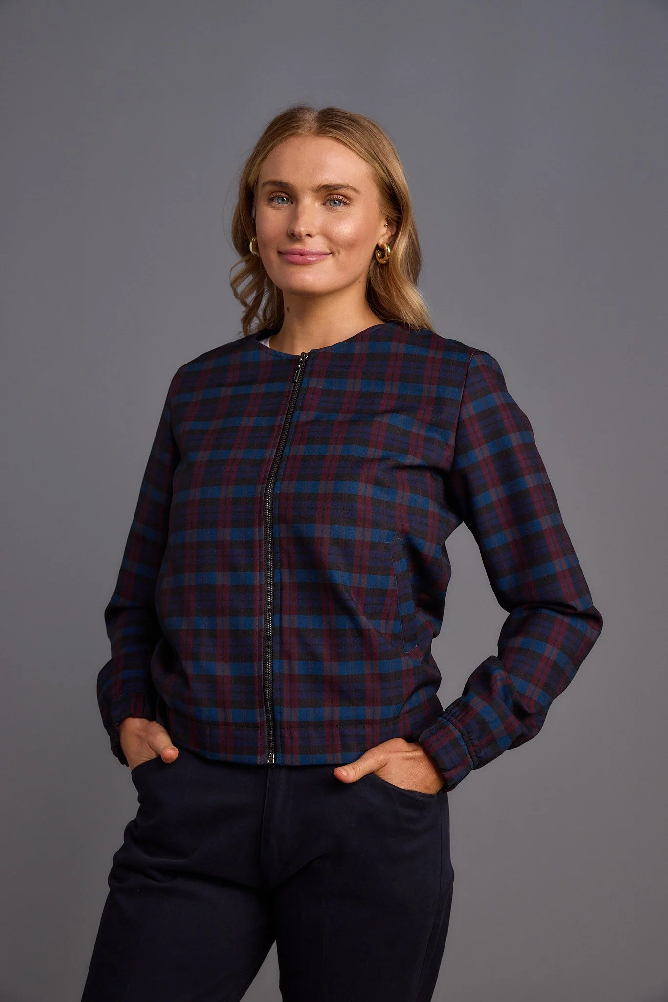 Woman with blonde hair wearing a checkered, zip-up jacket and dark pants, standing against a gray background.