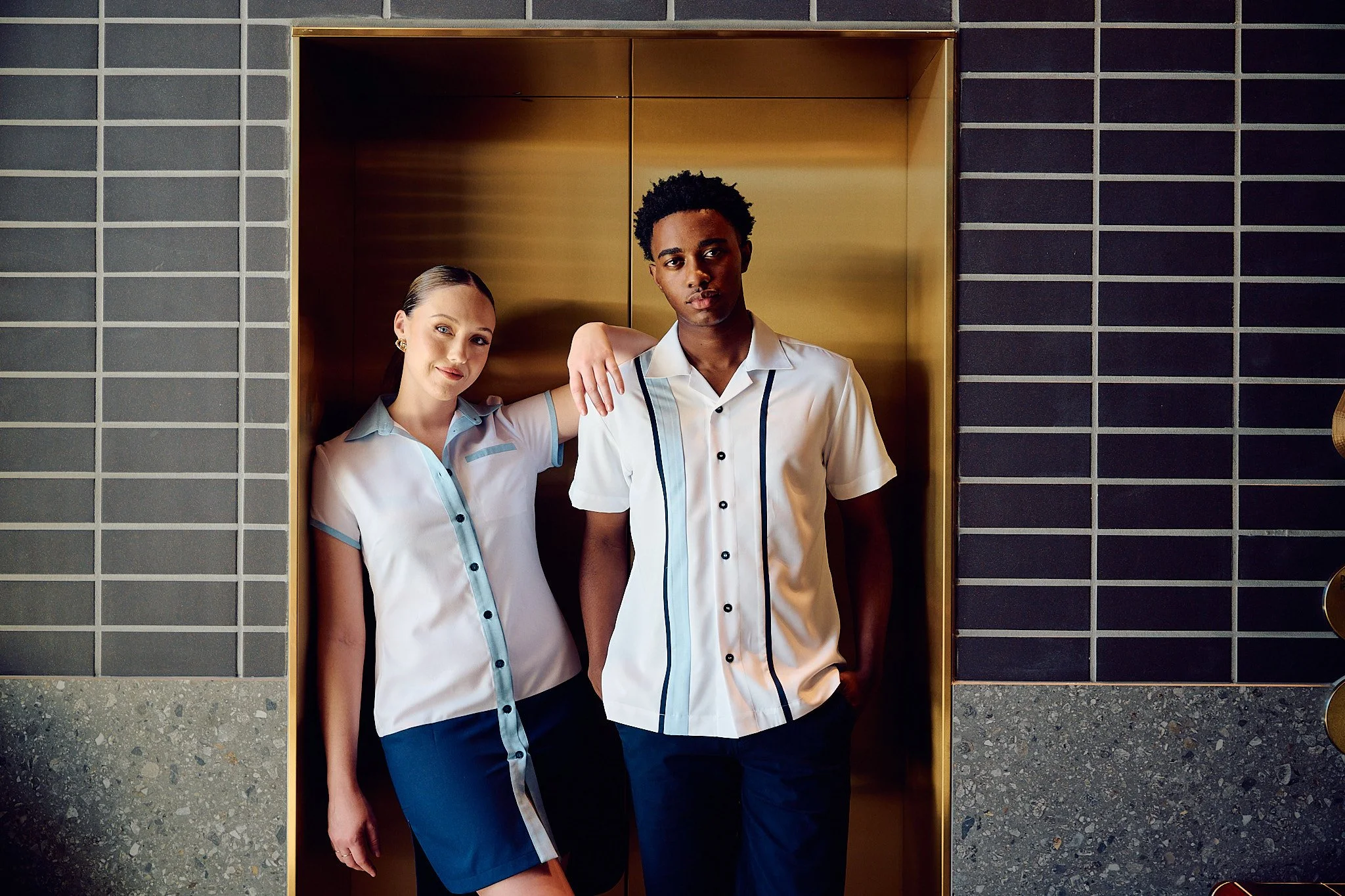 Two employees, a woman with blonde hair and a man with curly black hair, standing inside an elevator with gold doors. They are wearing matching outfits with light blue accents.