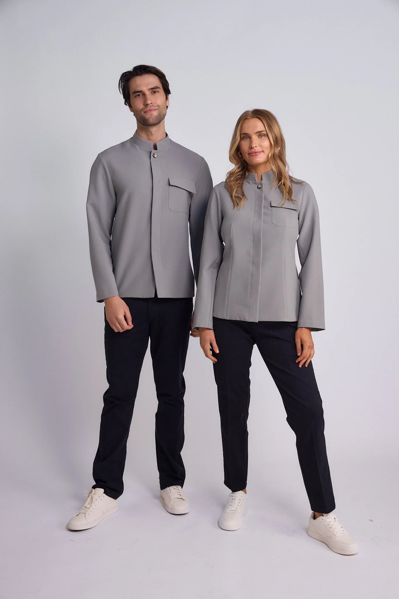 Two people, a man and a woman, standing side by side wearing matching light gray jackets and black pants, against a plain white background.