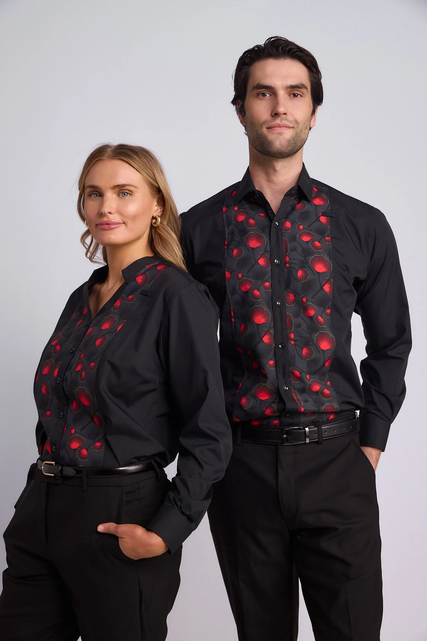 A man and a woman standing side by side, both wearing black shirts with red floral patterns and black pants, against a plain gray background.