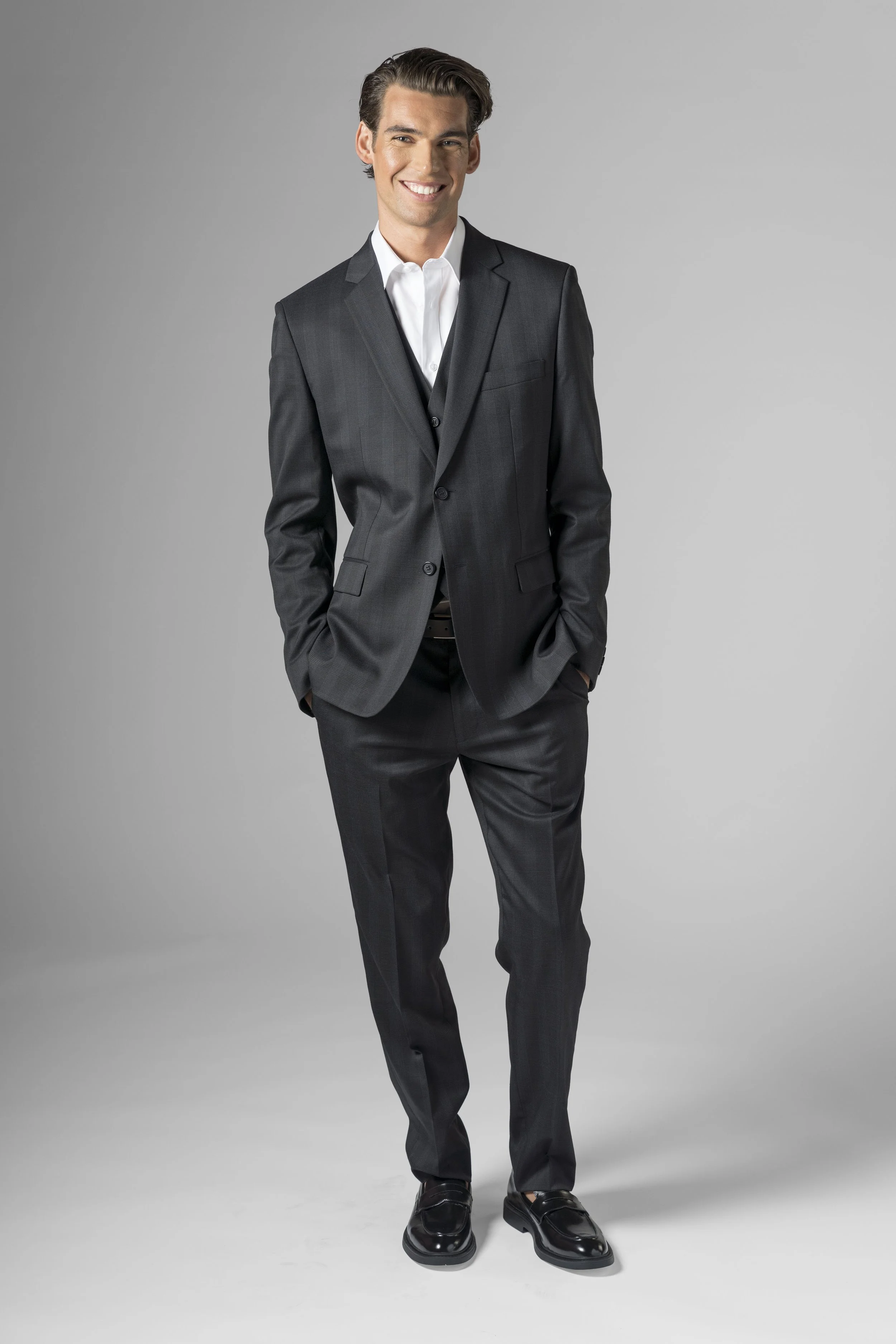 A young man with dark hair wearing a dark gray suit, white shirt, and black shoes, smiling and standing with hands in pockets against a plain light gray background.