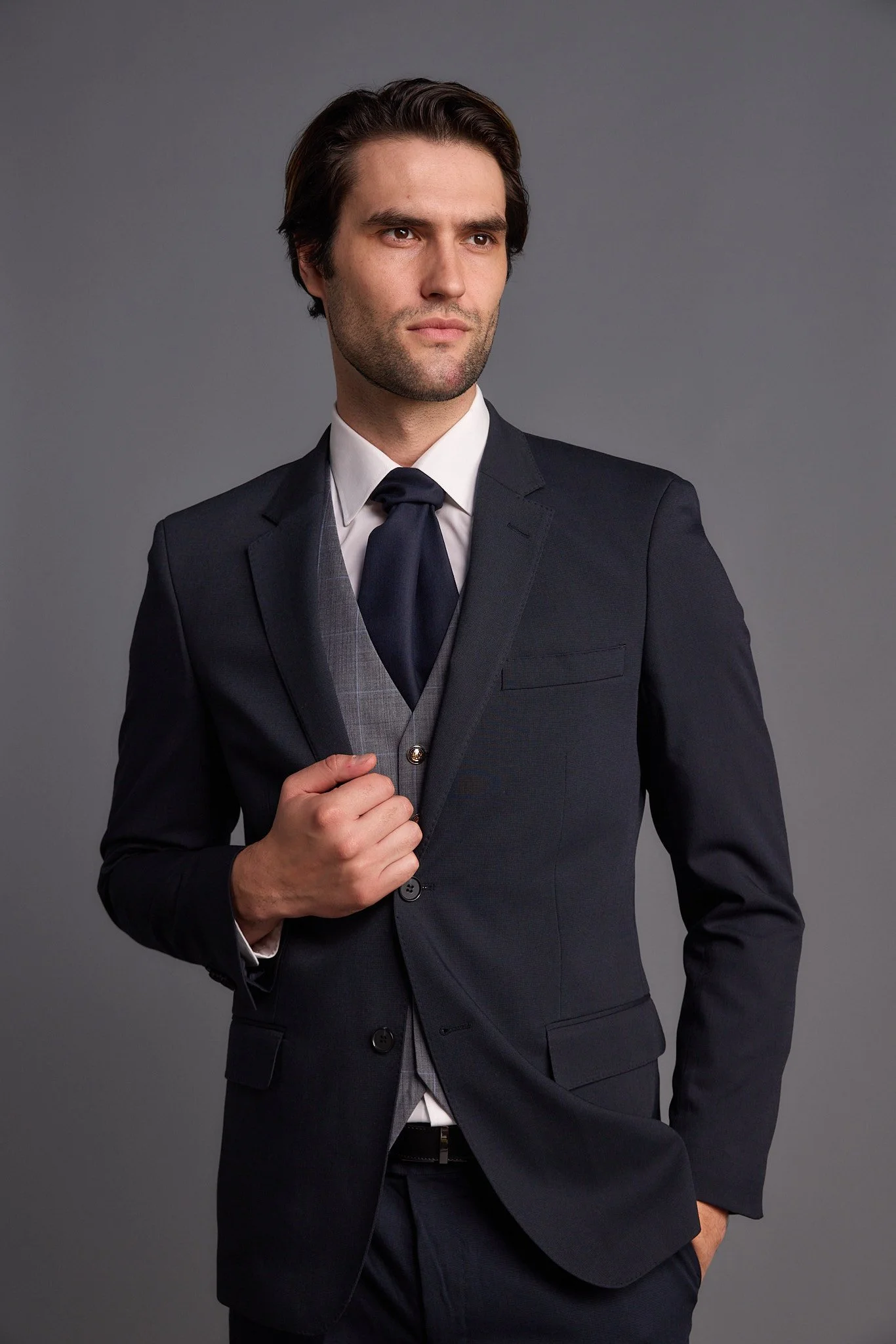 A man wearing a dark suit, white shirt, and tie, standing against a plain background.