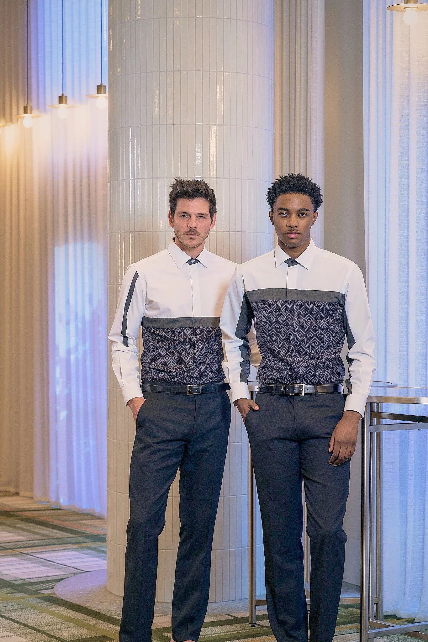 Two men wearing matching patterned shirts and dark pants standing in a well-lit, modern interior space.