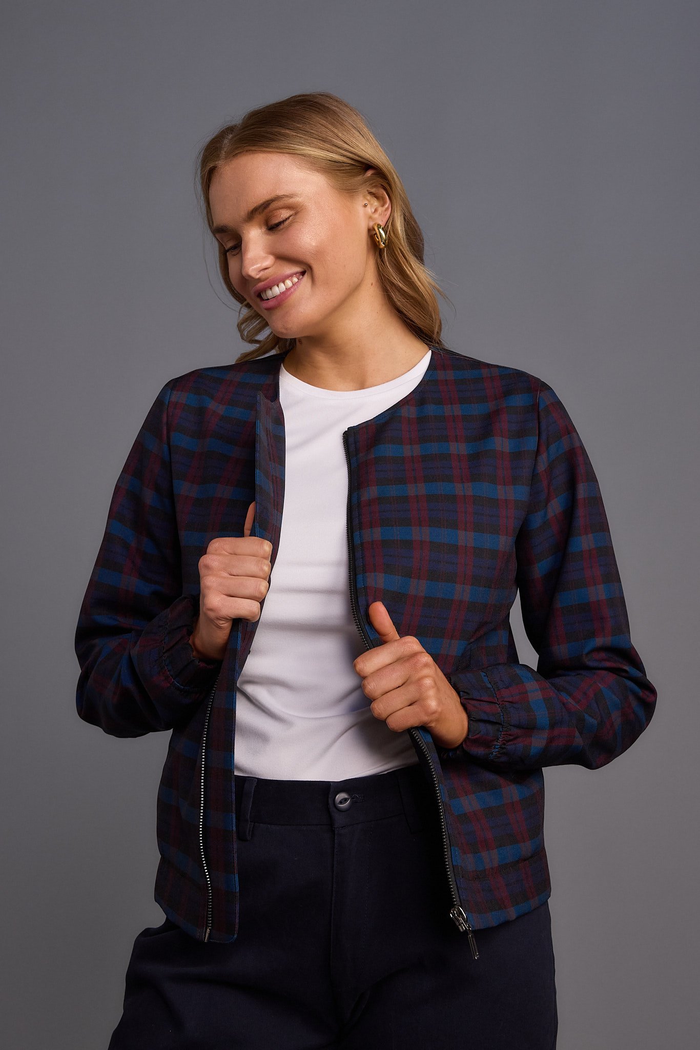 A woman with blonde hair wearing a plaid jacket, white t-shirt, and black pants, smiling with eyes closed against a gray background.
