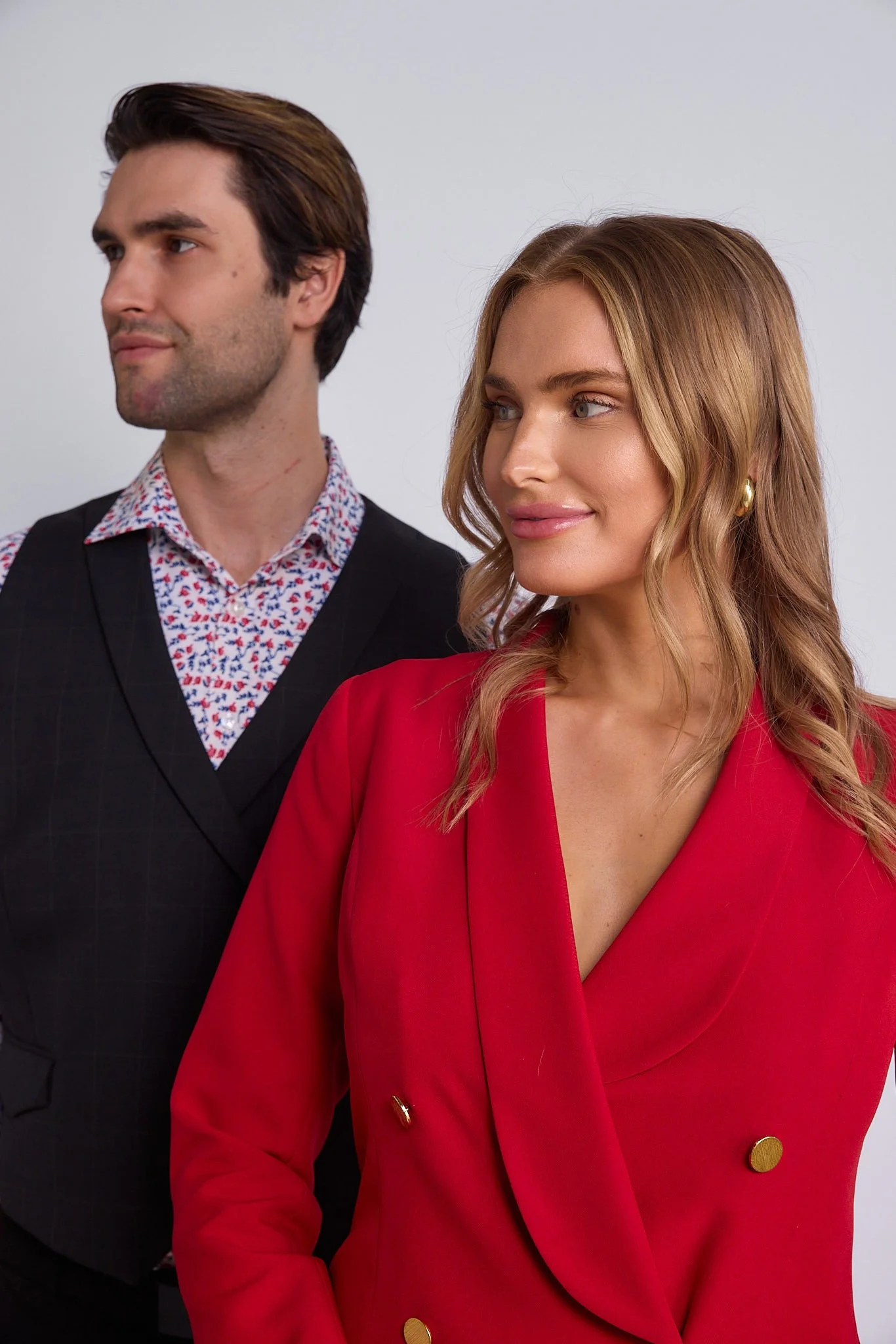 A man with dark hair in a black vest and a woman with wavy blonde hair in a red blazer, both looking to the side against a plain background.